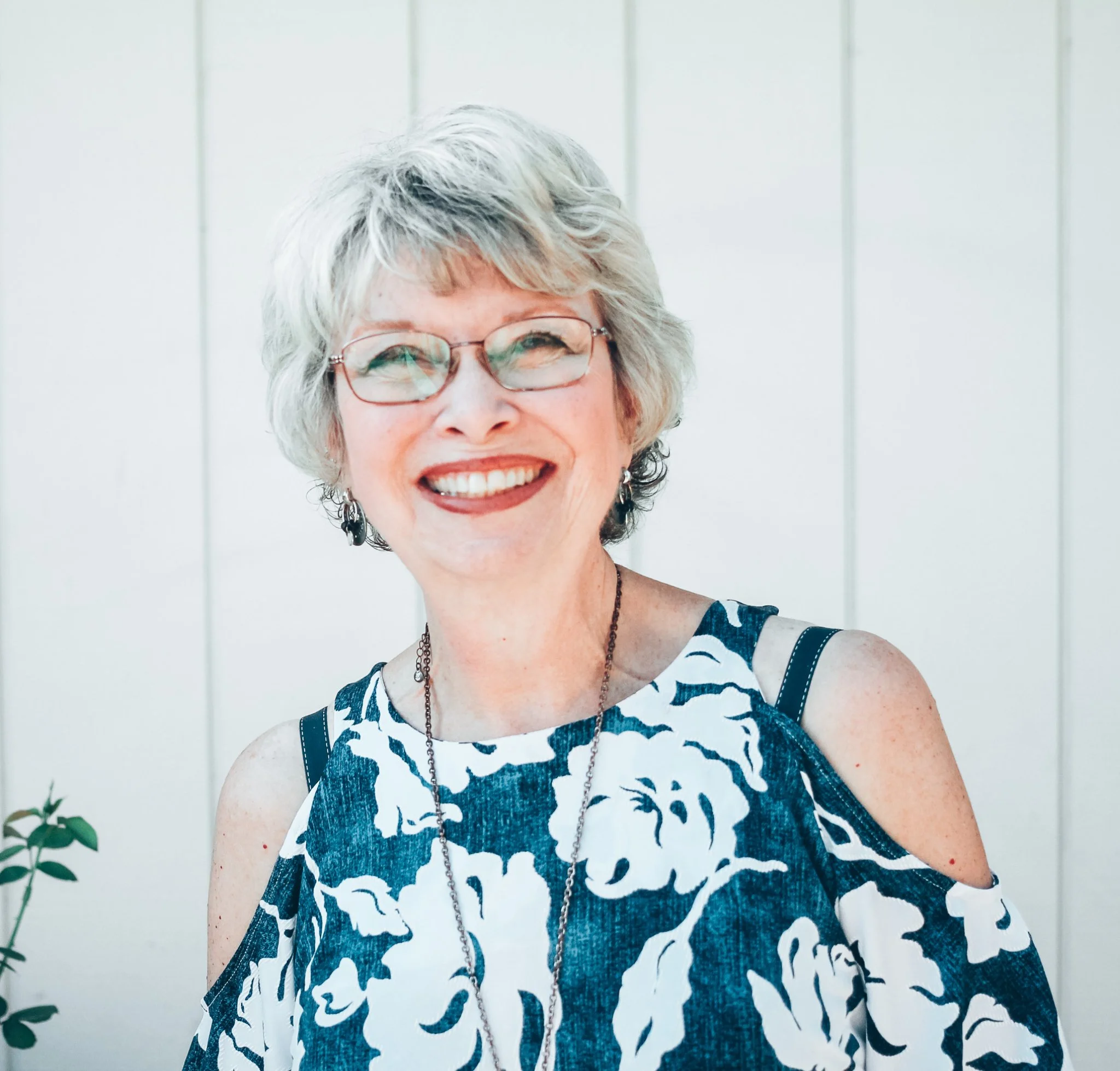 Smiling elderly woman with short gray hair, glasses, and earrings, wearing a blue and white floral cold-shoulder dress in front of a light-colored wall.