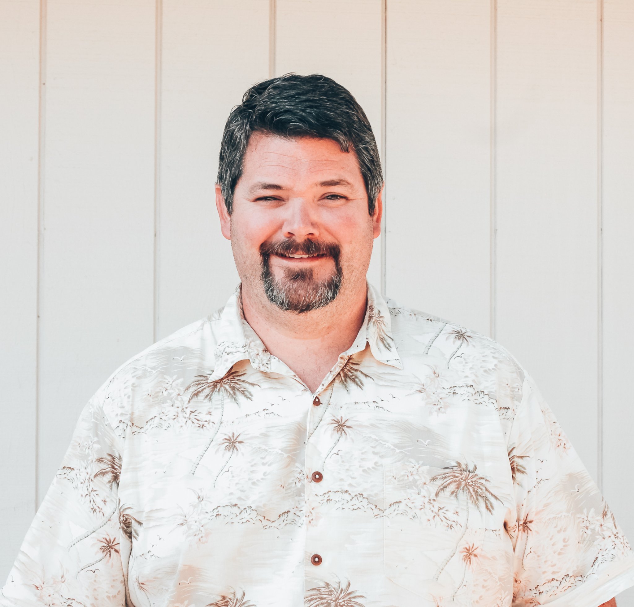 Man with dark hair and beard smiling, wearing a beige Hawaiian shirt with palm tree and island print, standing in front of a light-colored wooden wall.