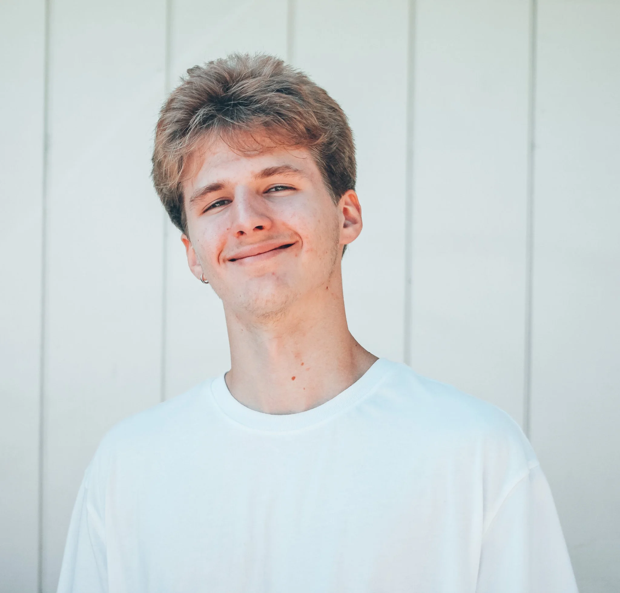 A young man with light skin, short wavy brown hair, and a small earring in his left ear, smiling with slightly squinting eyes, wearing a plain white t-shirt, standing in front of a light-colored vertical wooden panel background.