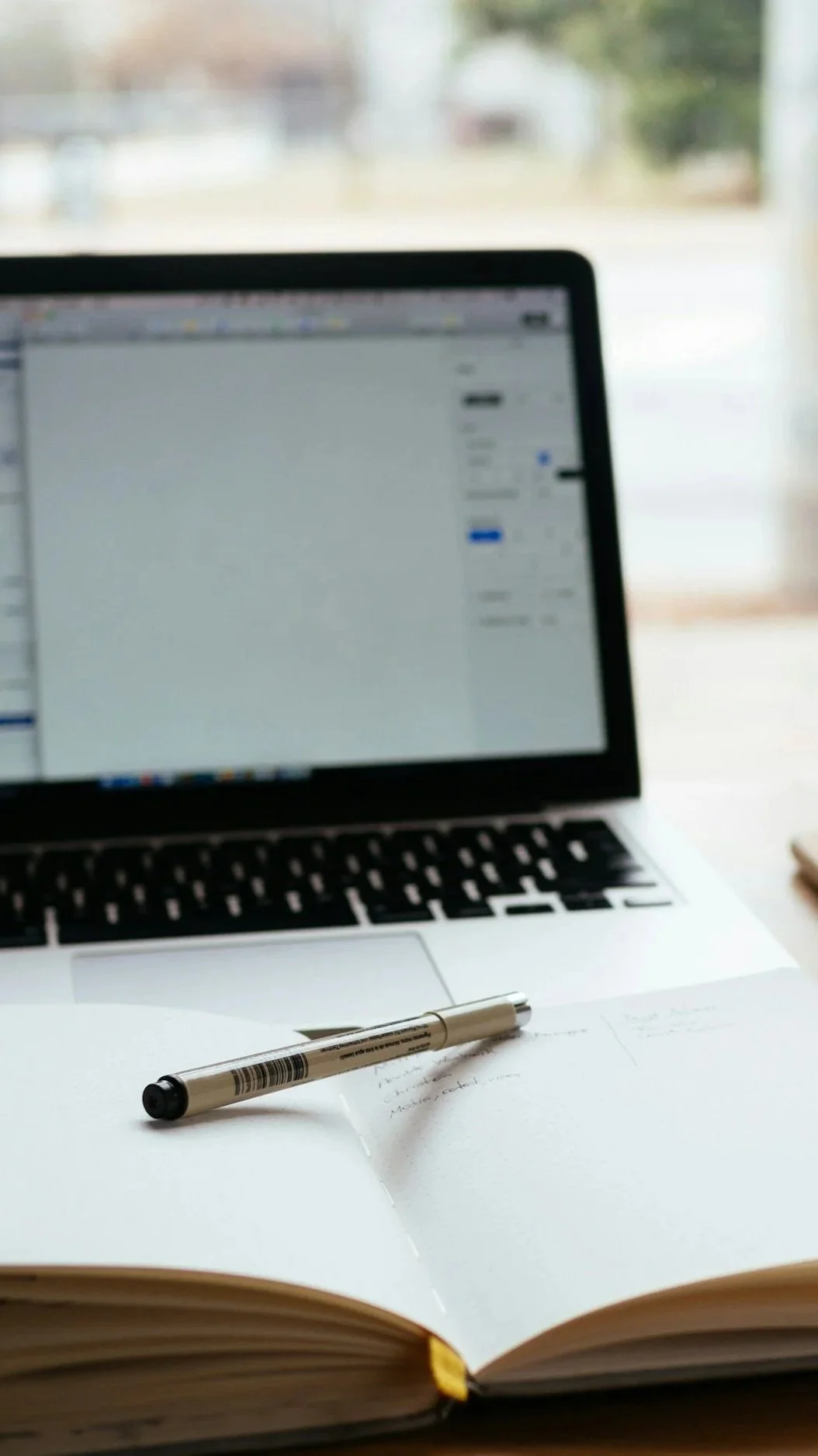 Open notebook with handwritten notes and a silver pen on top, in front of a laptop with a blurred screen, set near a window.