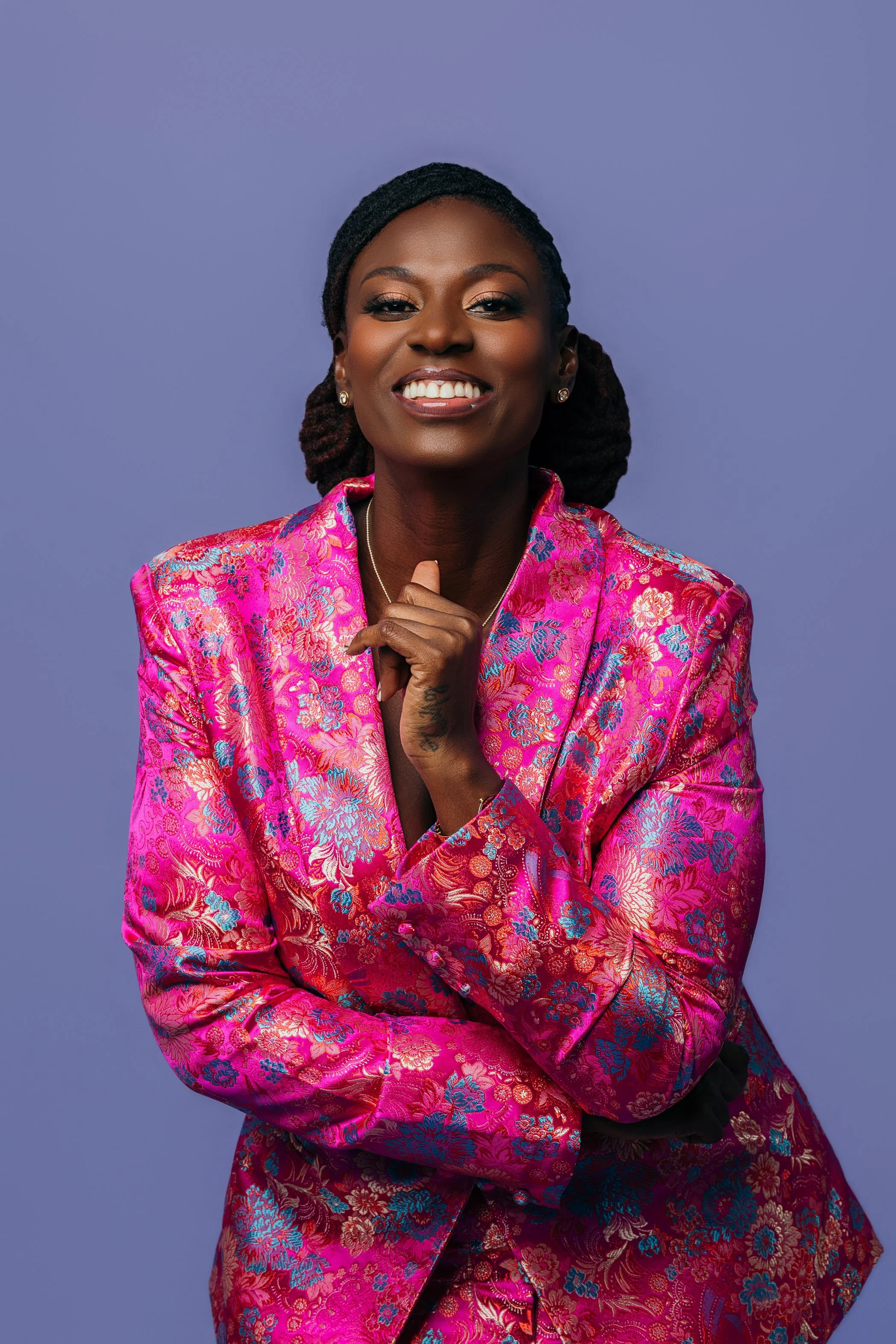 A woman with dark skin and braided hair, smiling, wearing a vibrant pink floral patterned blazer, against a light purple background.