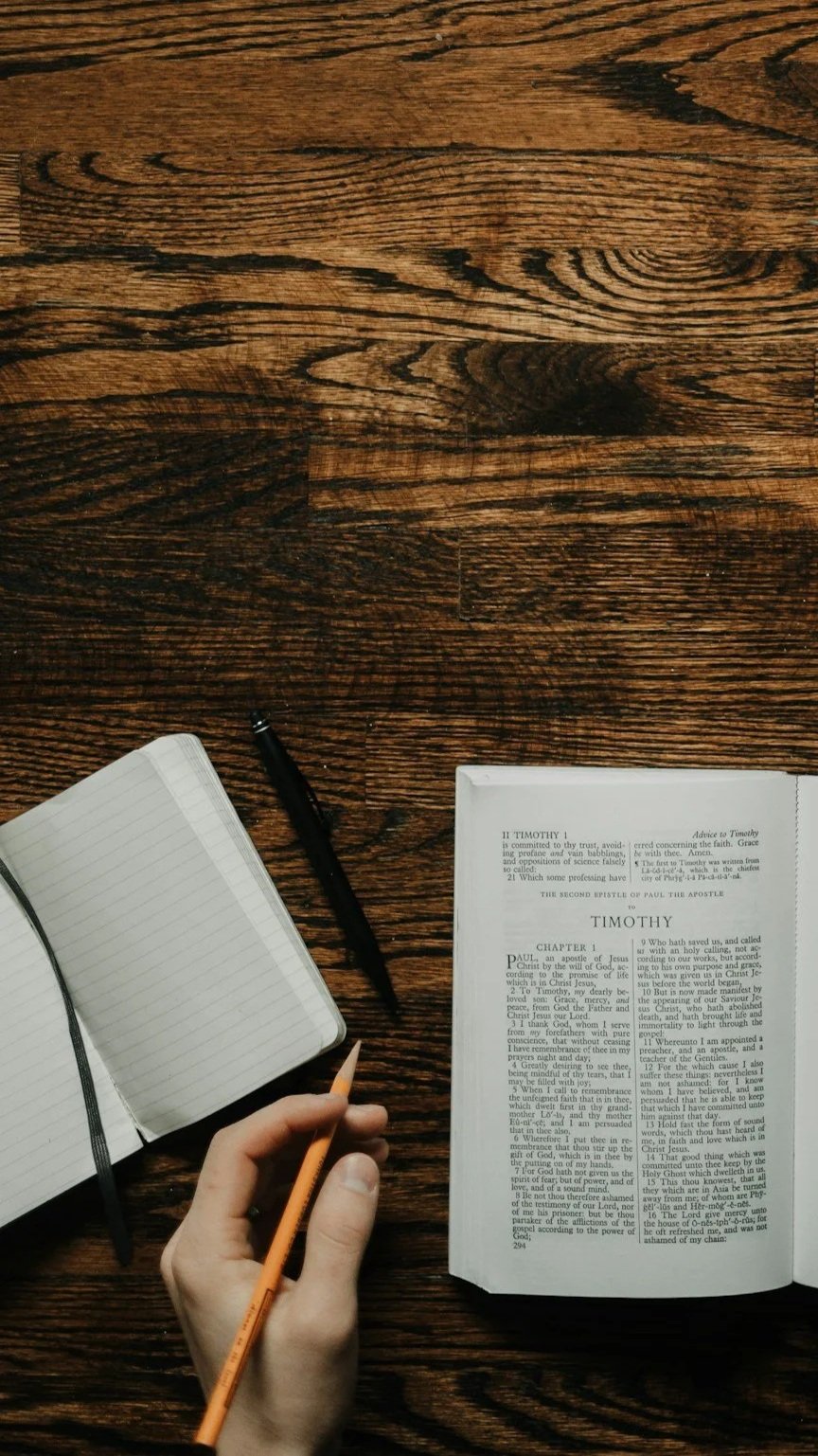 Open Bible and notebook on a wooden table, person holds a yellow pencil.