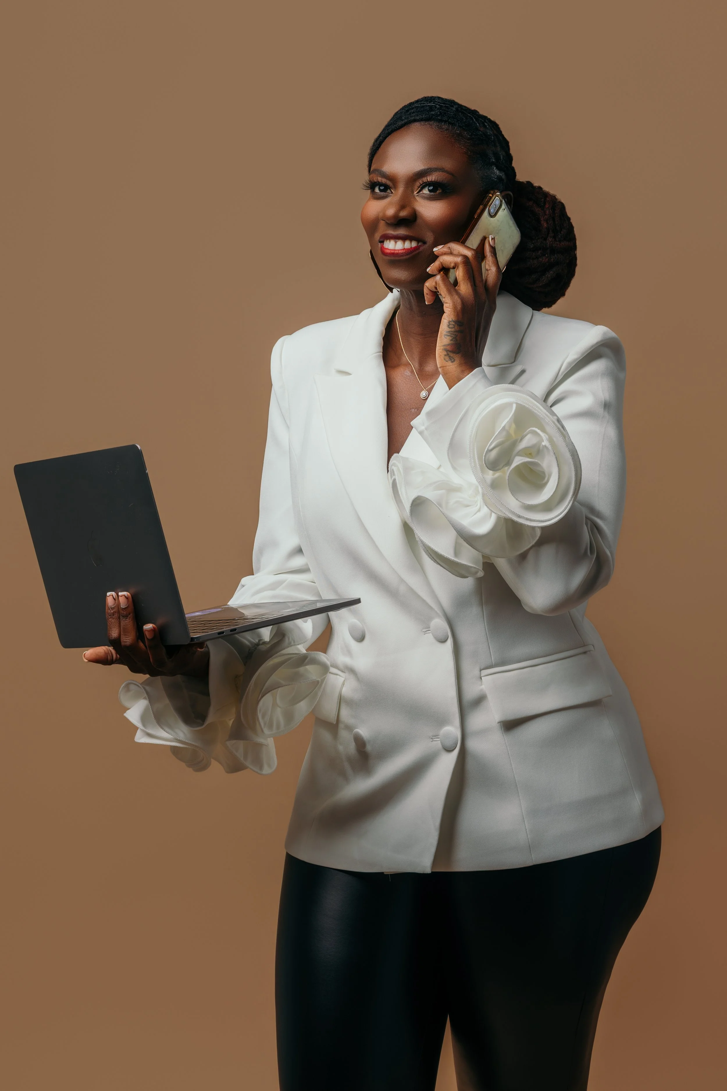A woman with dark hair styled in braids, wearing a white blazer with ruffled sleeves, holding a laptop in one hand and talking on a cell phone, standing against a beige background.