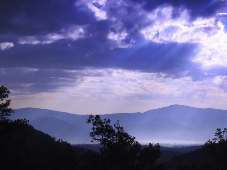A photo of Shenandoah valley with a mountain landscape with dark clouds and blue sky, silhouetted trees in the foreground.