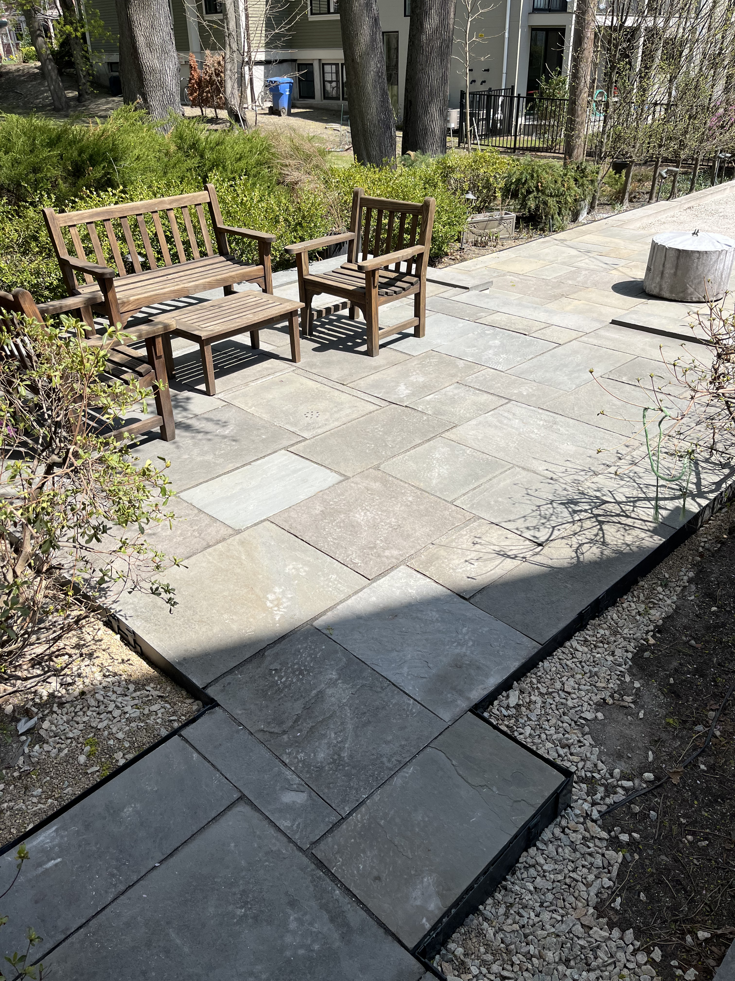 Outdoor patio area with two wooden benches and a small wooden table, surrounded by bushes and trees, adjacent to a gravel walkway, with residential buildings in the background.
