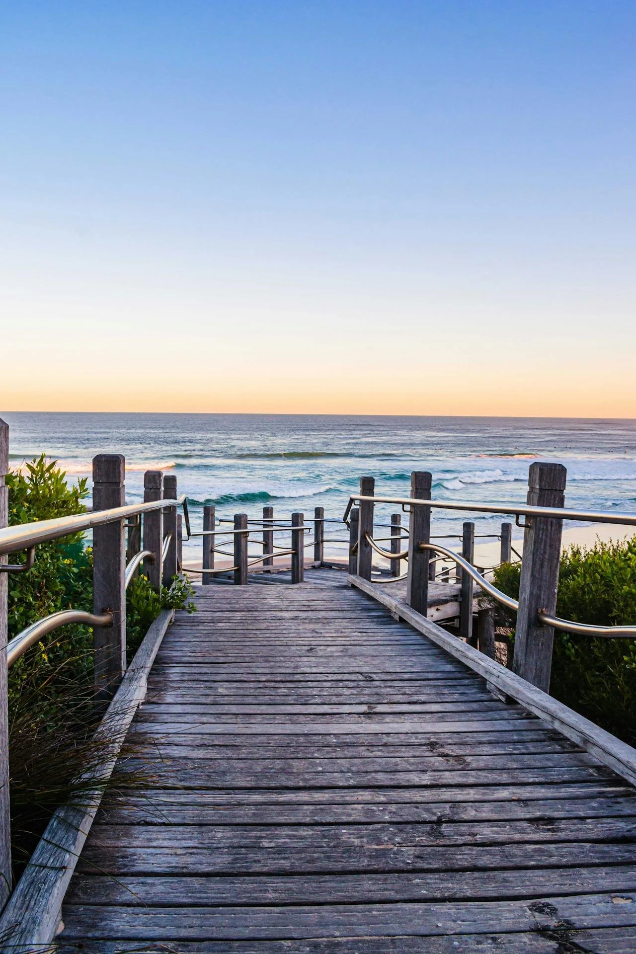 Wooden walkway leading to the beach with ocean waves and a sunset sky in the background.