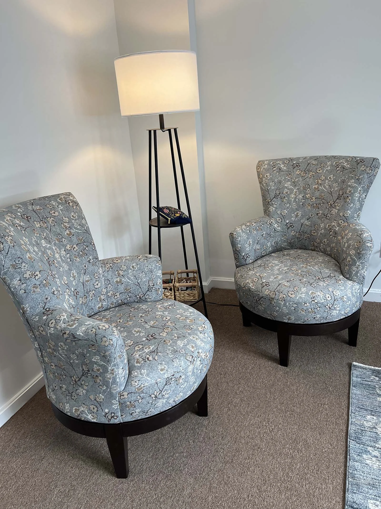 Two upholstered armchairs with floral fabric pattern, placed on a brown carpeted floor, next to a black floor lamp with a white lampshade in a corner of a room.