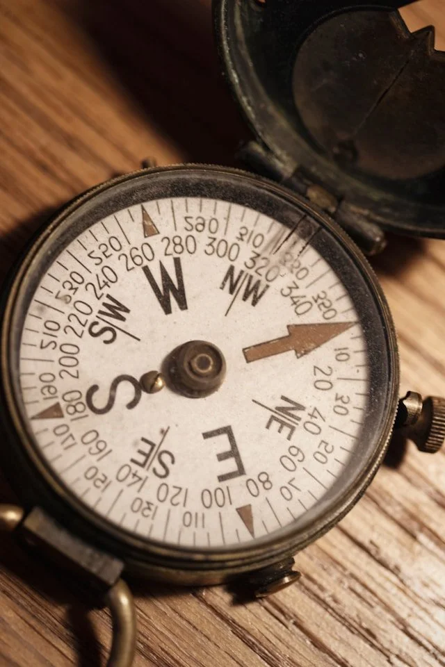 A vintage compass showing west at approximately 250 degrees, placed on a wooden surface with a small open case nearby.