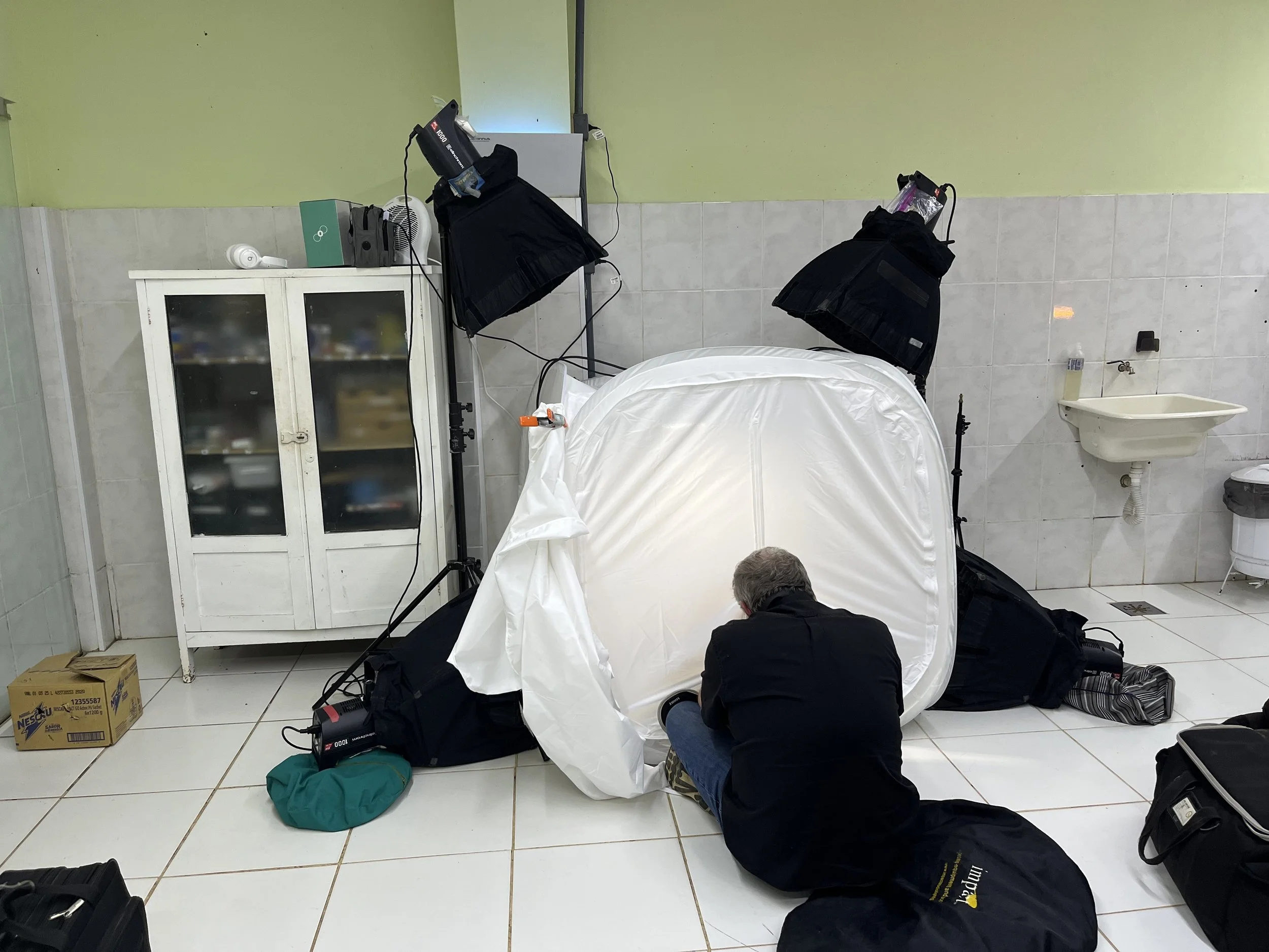 Joel Sartore sitting on the floor in front of a white photography light tent with two large studio lights overhead, in a room with tiled floor and walls, and a small sink on the wall during a Photo Ark photography session in Brazil.