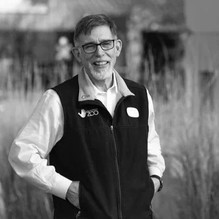 John Chapo smiling with glasses, wearing a vest and white shirt, standing outdoors with a blurred background as co-treasurer and board member of the Photo Ark Foundation