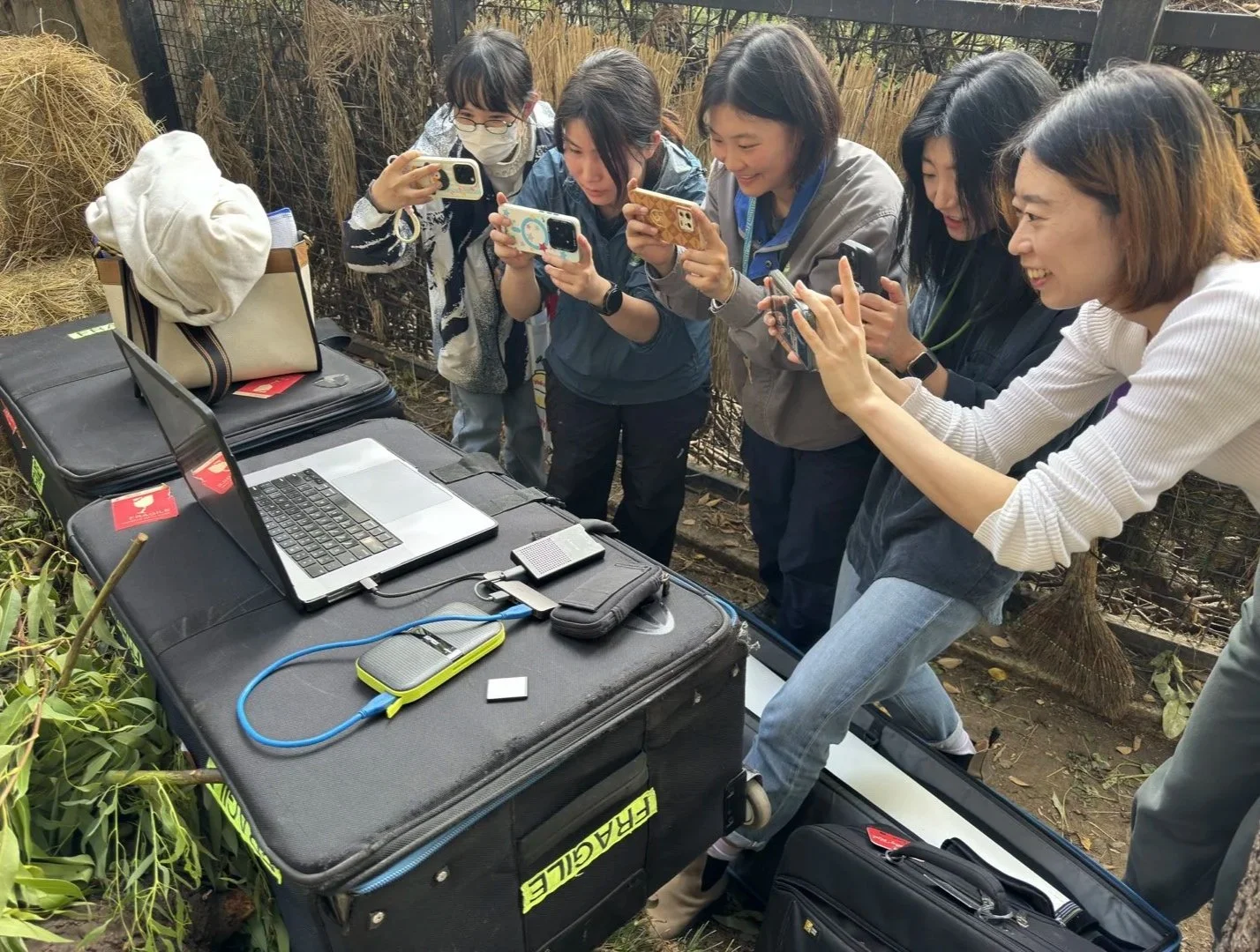 Six women are gathered around a table outdoors, taking photos or videos with their smartphones, with a laptop and electronic equipment on the table.