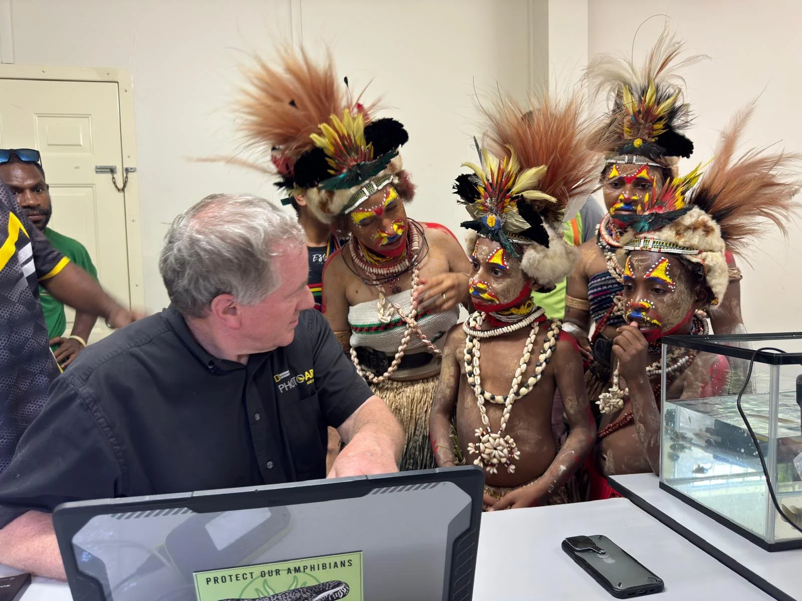 Joel Sartore in a black National Geographic Photo Ark shirt sits at a table with young children and adults in traditional tribal attire, face paint, and feathered headdresses in Port Moresby, Papua New Guinea, during an educational event.