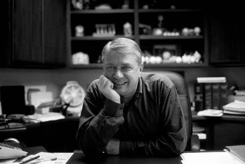 Kyle Sitzman sitting at a cluttered desk in a home office, smiling, with books and decorative items on shelves in the background as a co-treasurer and board member of the Photo Ark Foundation.