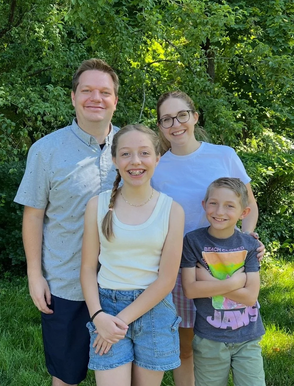 A family of five posing outdoors in front of green foliage, smiling at the camera.