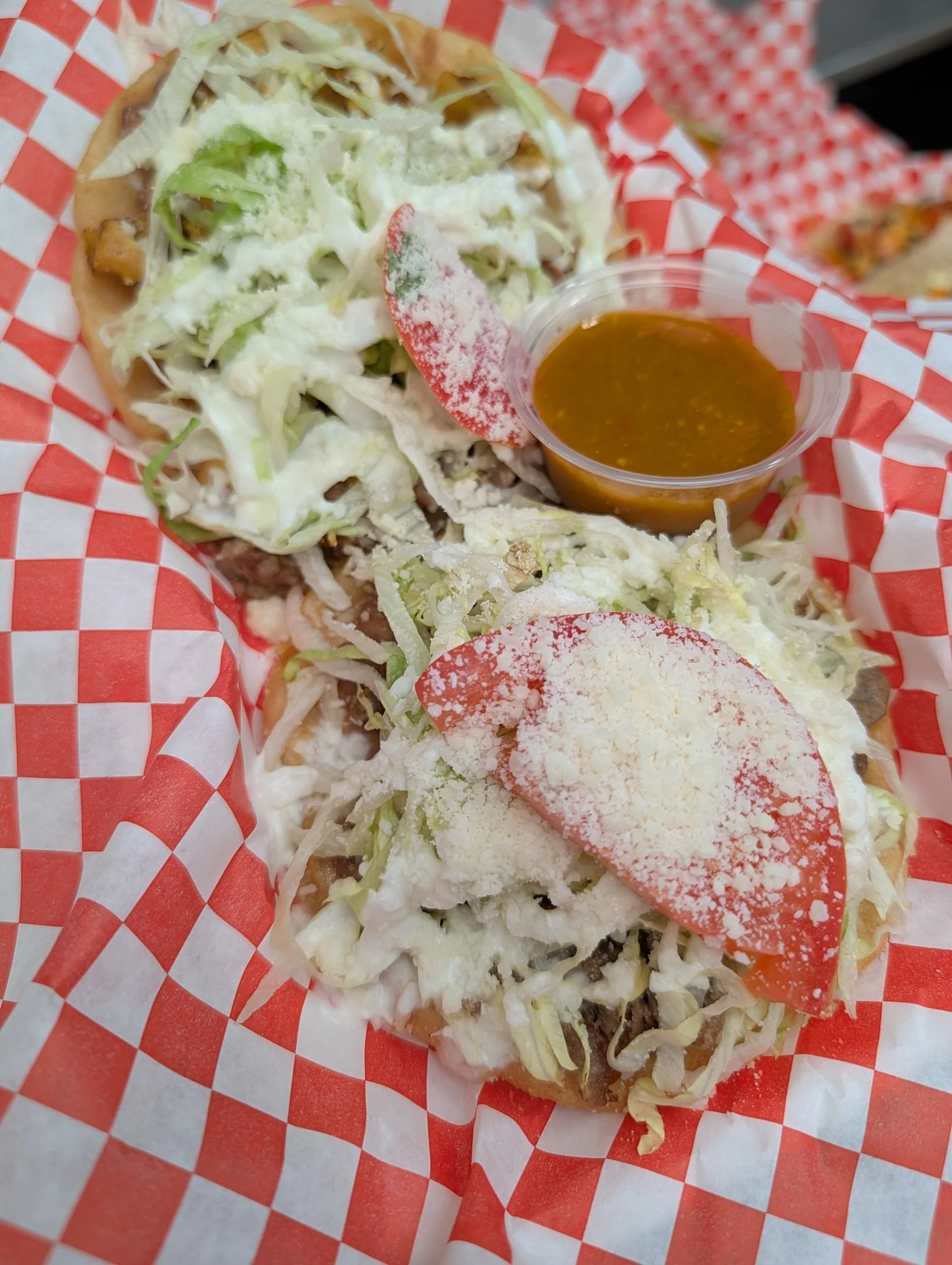 Two tacos filled with shredded lettuce, cheese, and meat, topped with tomato slices and grated cheese, with a small cup of salsa on a red and white checkered paper.