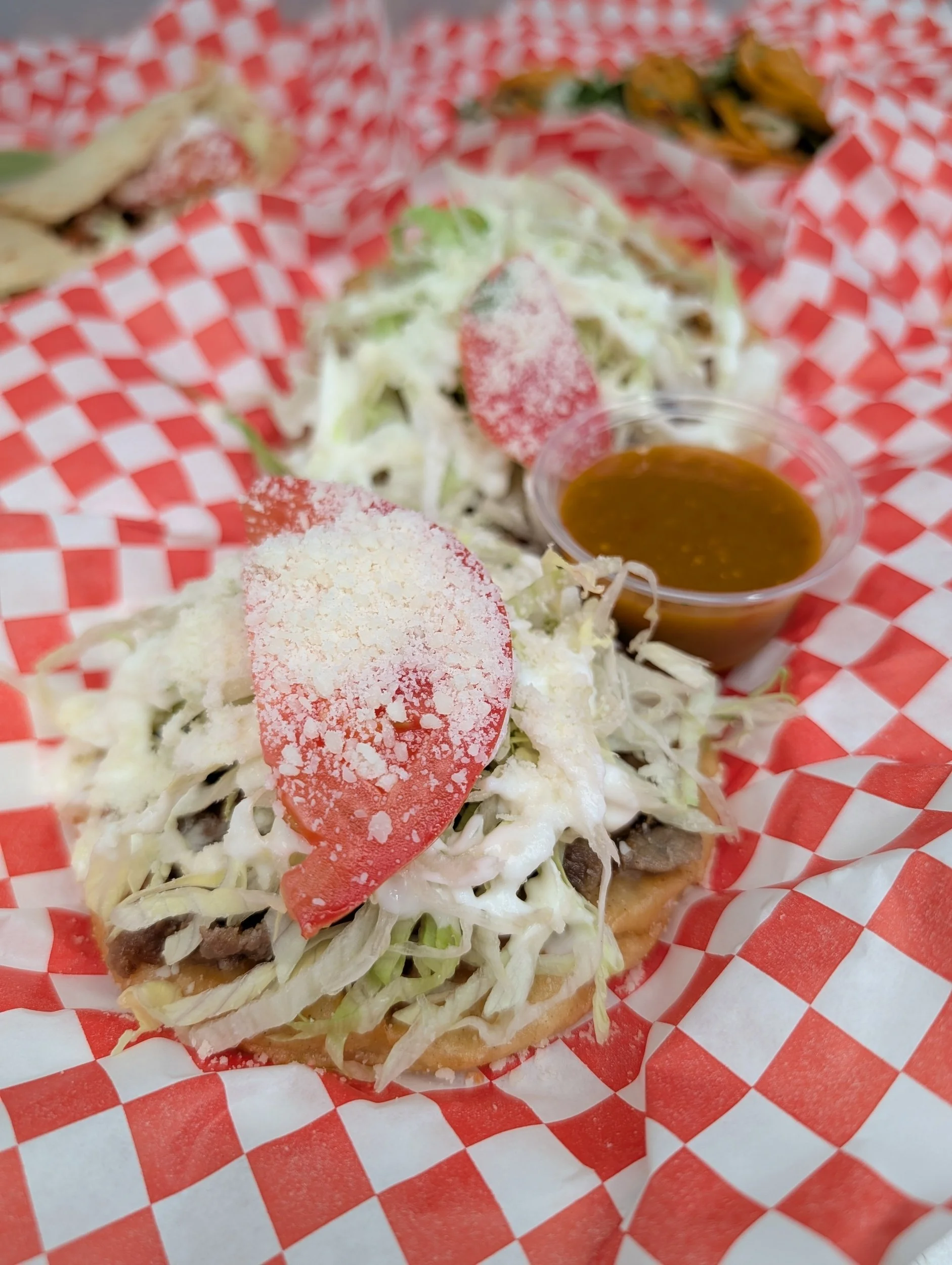 Two tacos with shredded lettuce, cheese, sliced tomatoes, and ground meat on a red and white checkered paper, with a small cup of brown sauce.