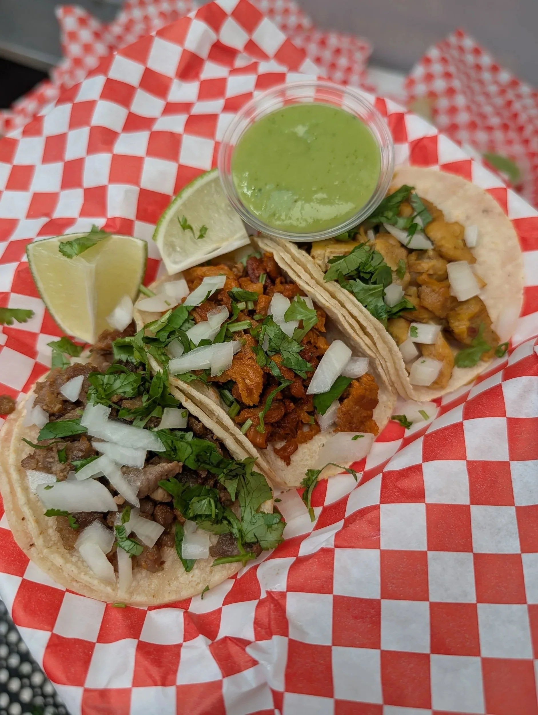 Three tacos on a red checkered paper with chopped onions and cilantro, a lime wedge, and a small container of green salsa.