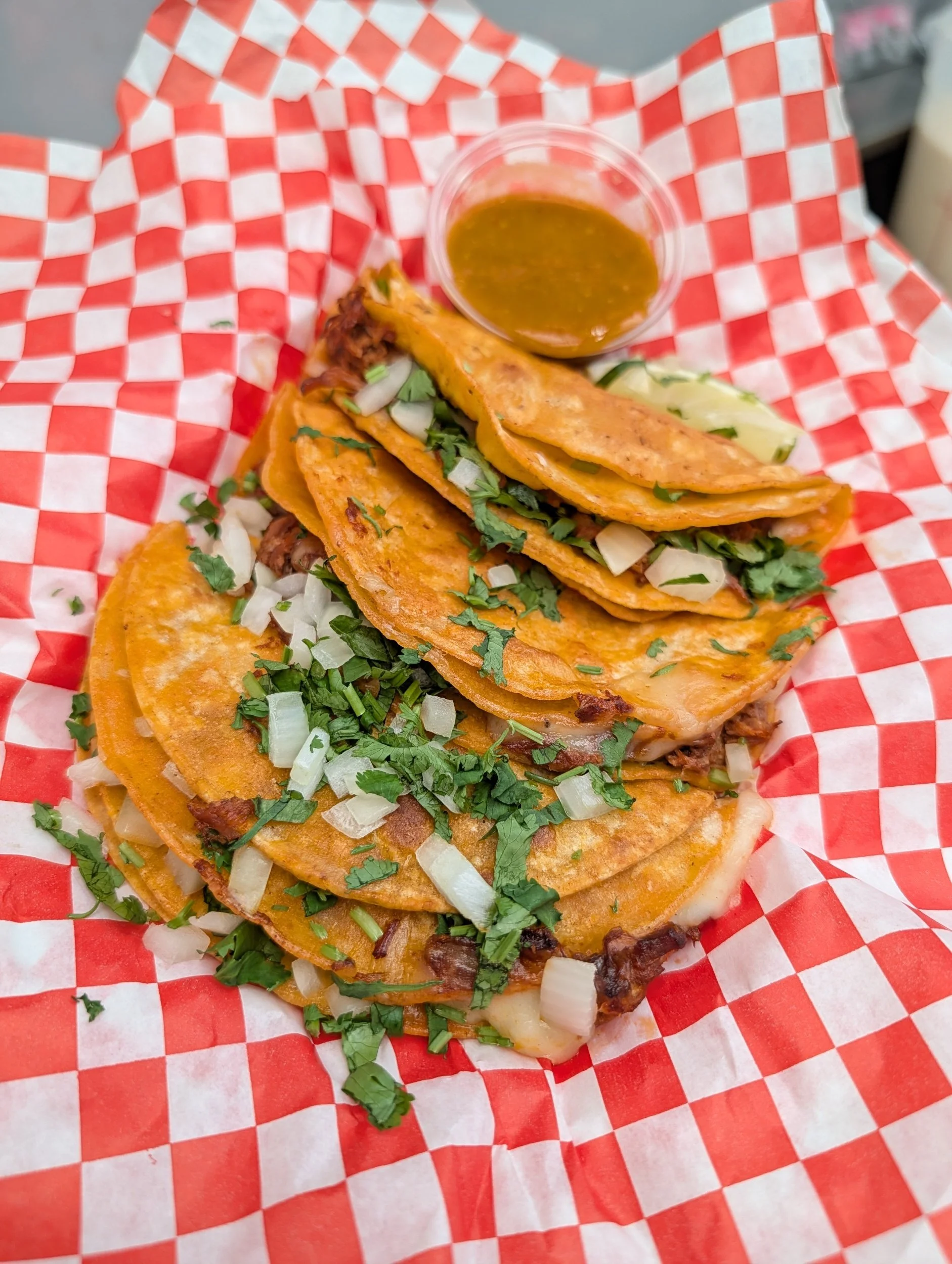 Three tacos filled with shredded meat, chopped onions, and fresh cilantro, served on red and white checkered paper with a small cup of sauce.