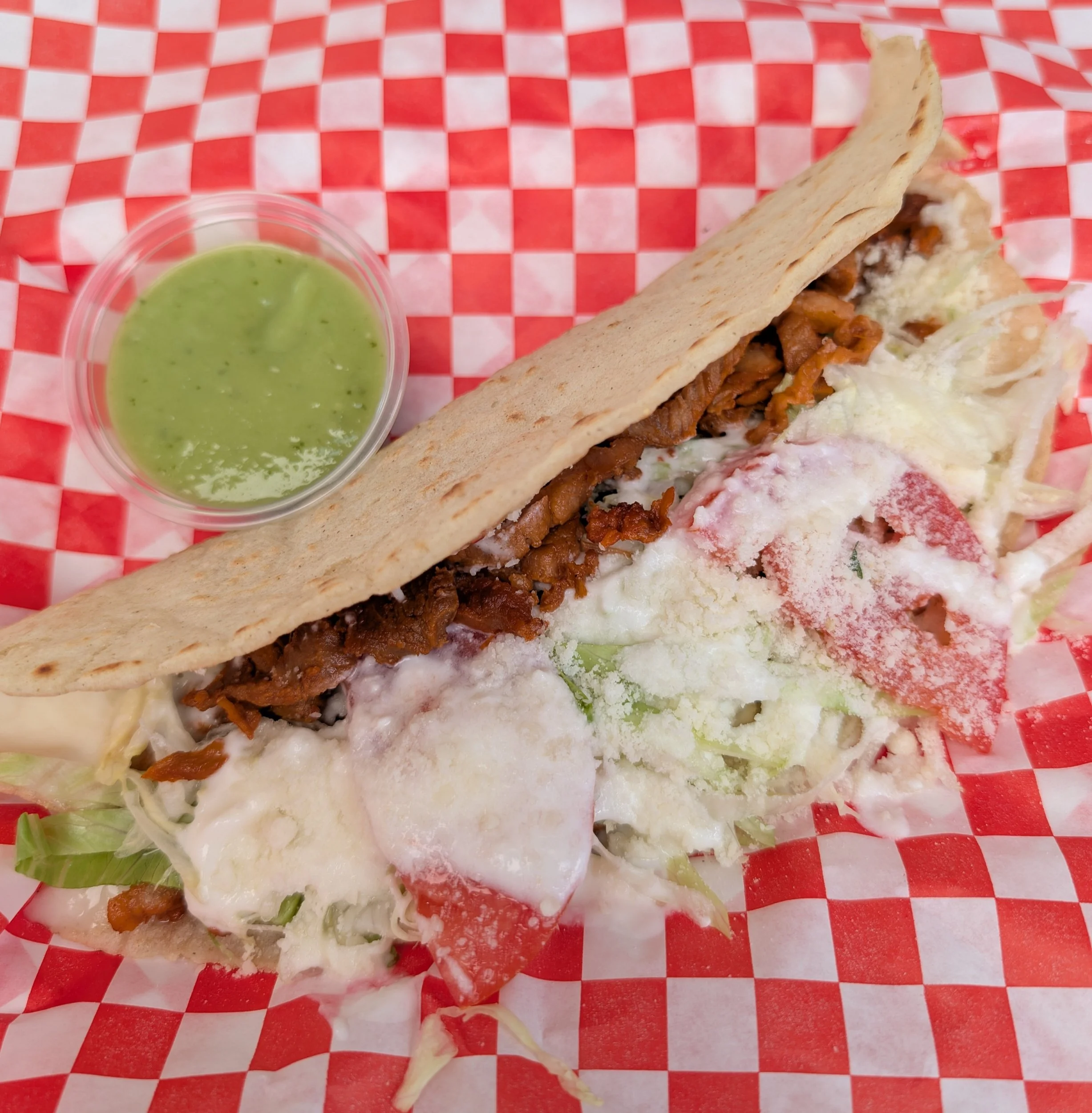 A taco with shredded beef, lettuce, tomatoes, cheese, and sour cream on a red checkered paper, served with a side of green sauce.