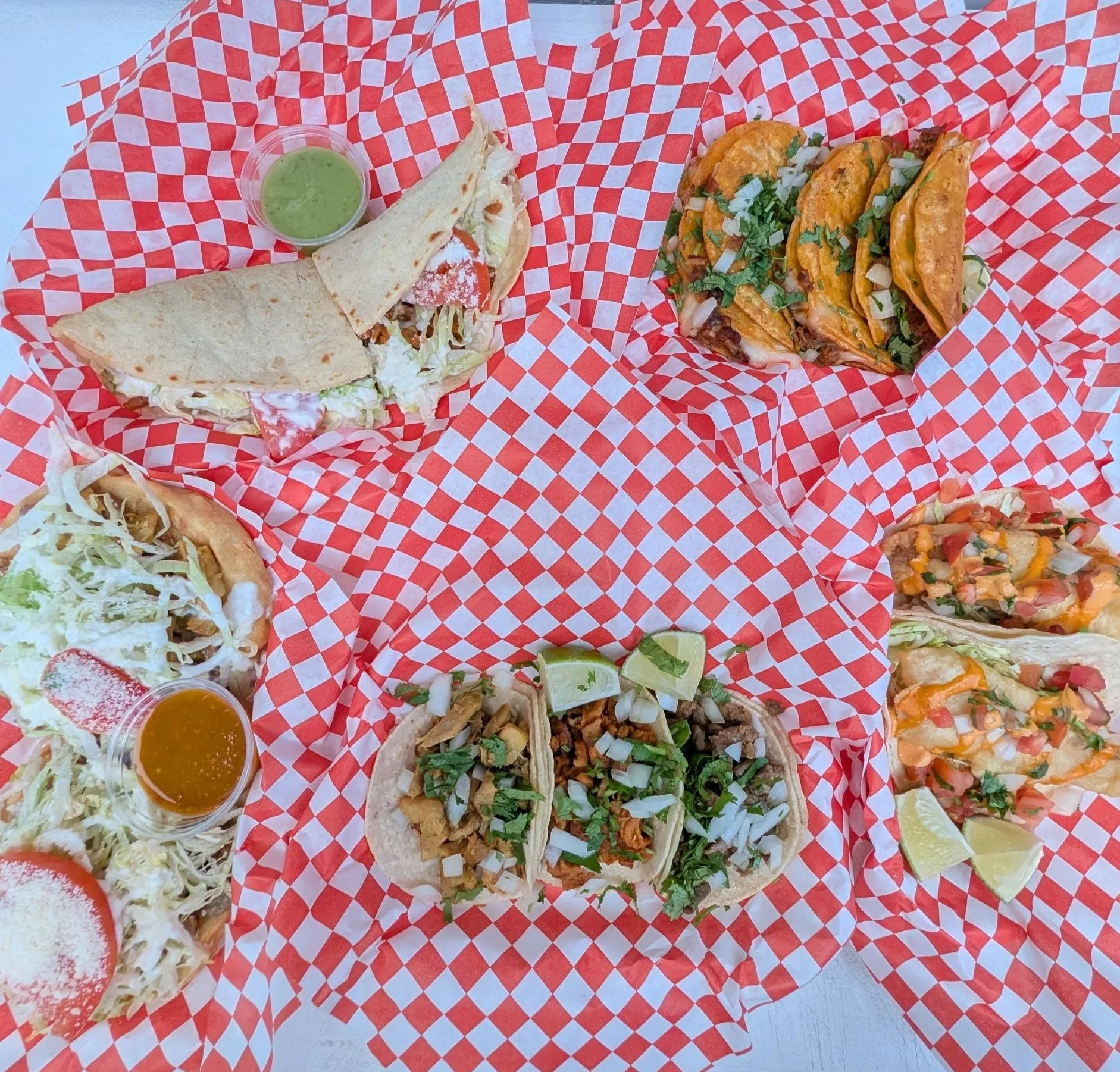 Four different types of tacos and a quesadilla on red checkered paper, with two small containers of dipping sauces and lime wedges.
