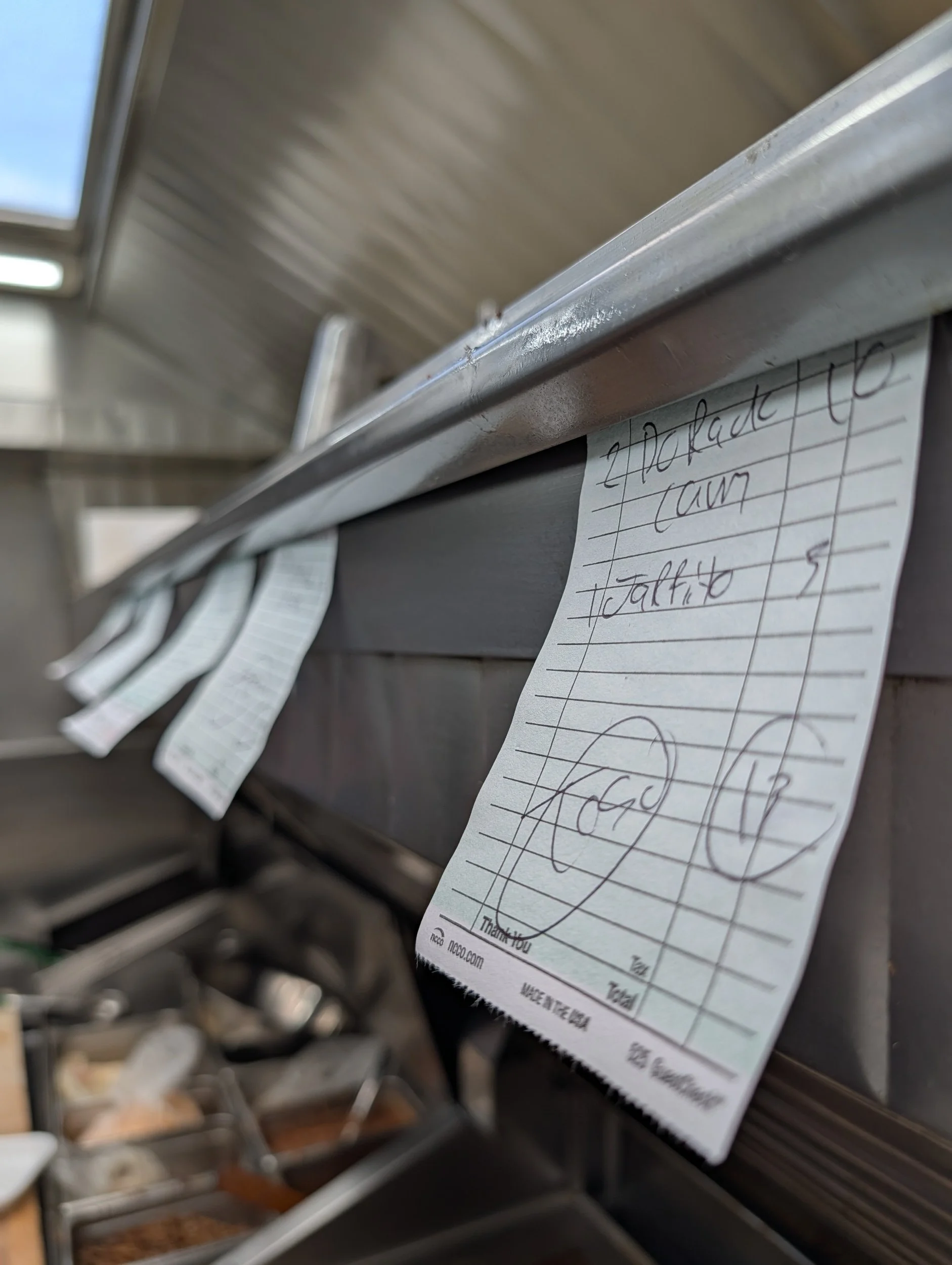A close-up of a handwritten order slip attached to a metal counter at a cafeteria or restaurant, with other slips and food containers visible in the background.