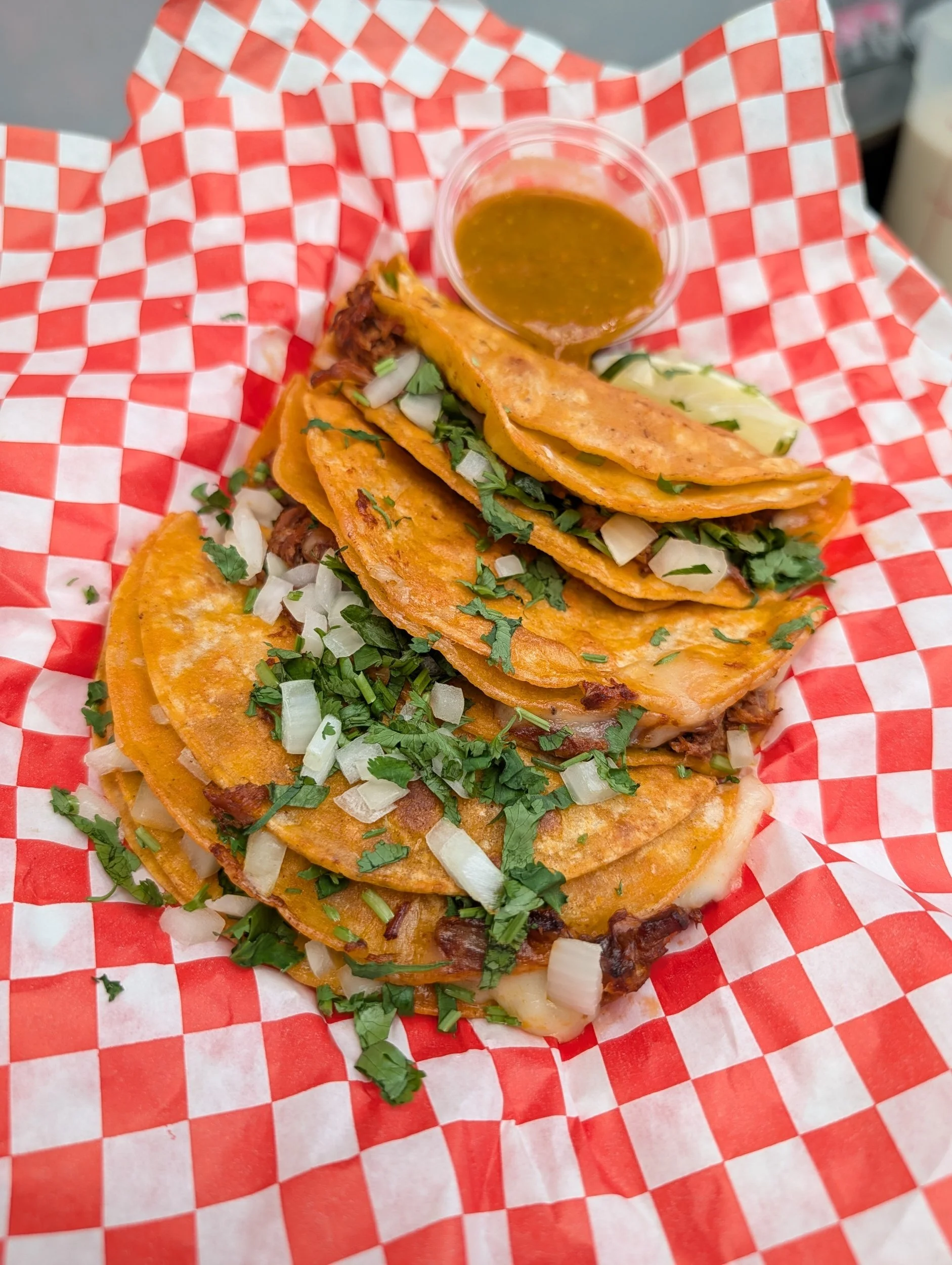 Three Mexican tacos with shredded lettuce, chopped onions, and cilantro, served with a small cup of hot sauce on red and white checkered paper.