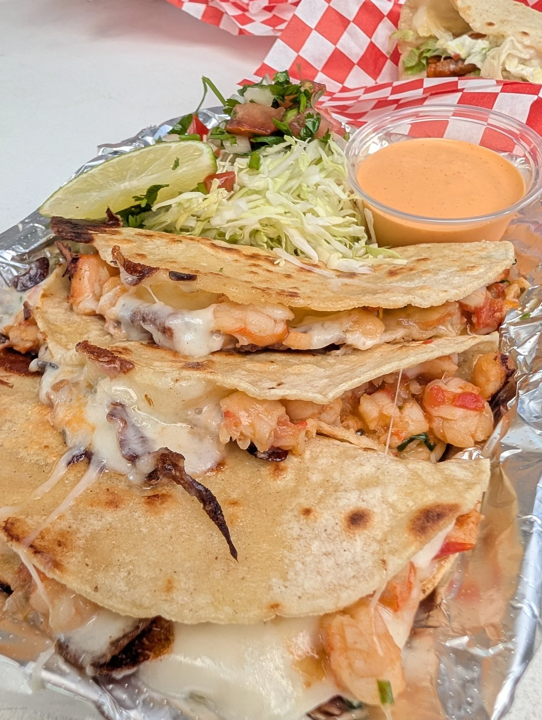 Three tacos with melted cheese, shrimp, and vegetables served with shredded lettuce, a lime wedge, a small container of orange sauce, and a side of shredded lettuce and pico de gallo on a foil-lined tray.