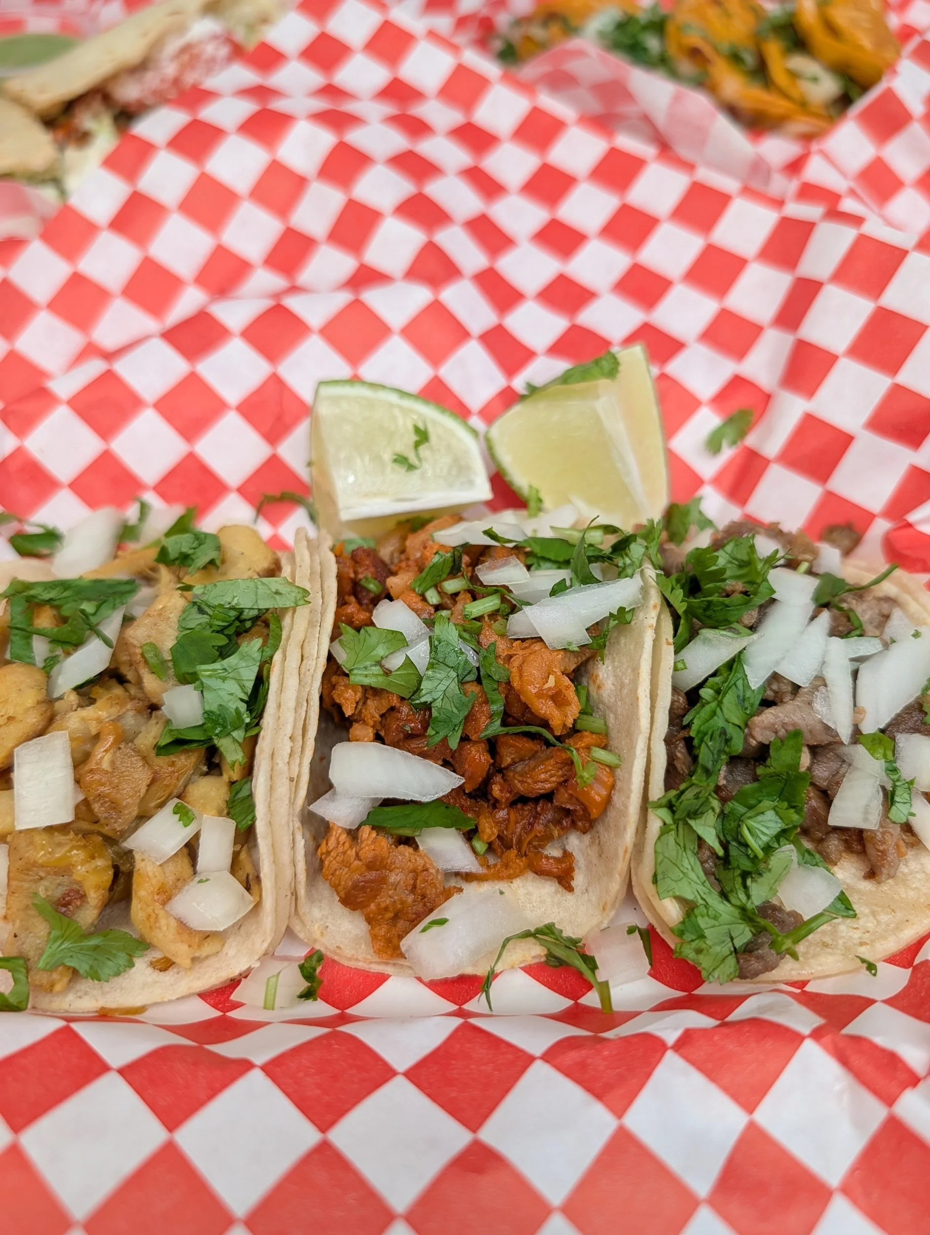 Three street tacos on a red and white checkered paper with lime wedges on top, garnished with chopped onions and cilantro.