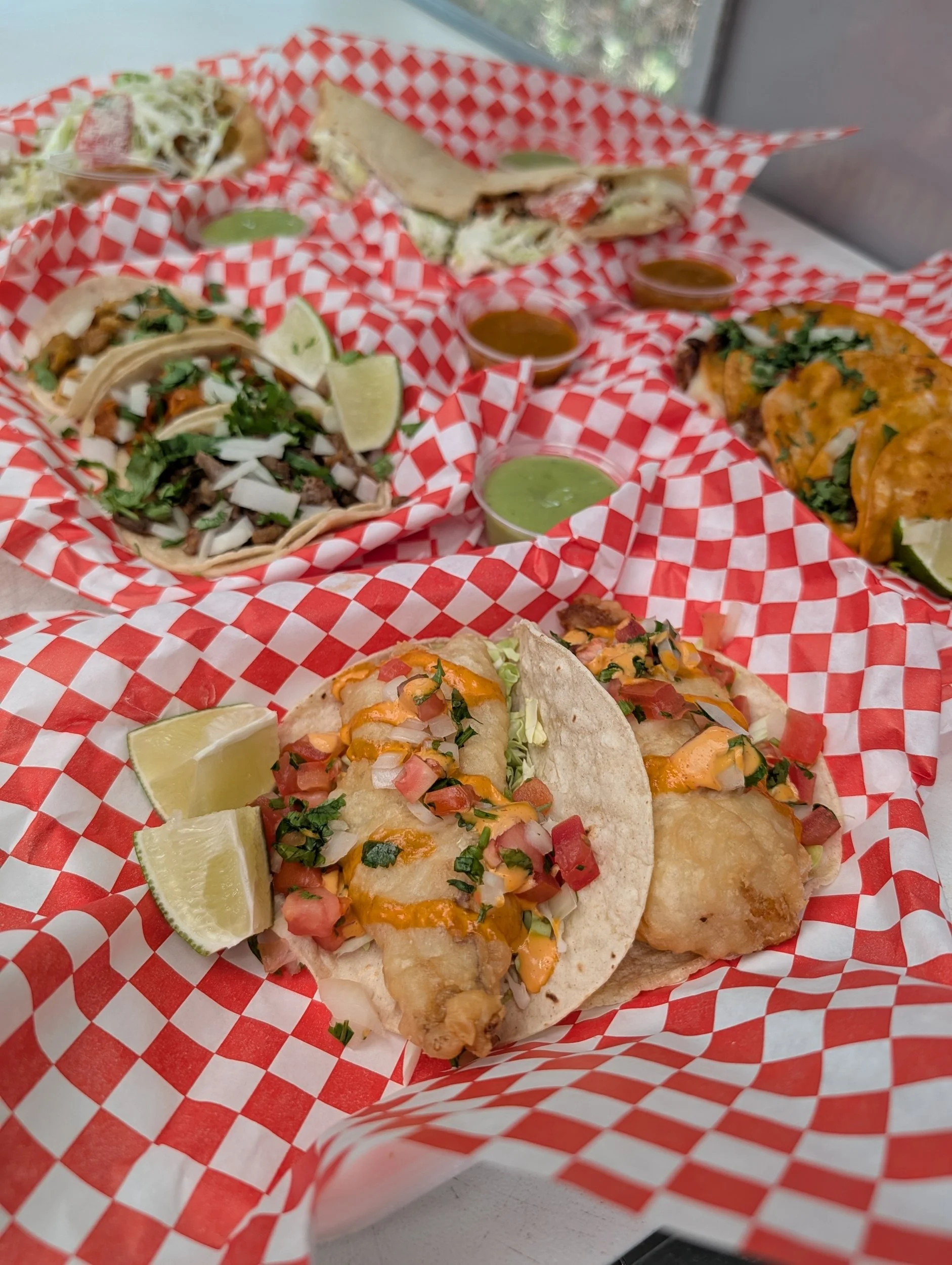 Three fish tacos with chopped onions, cilantro, and sauce, accompanied by lime wedges and green sauce, on red and white checkered paper.