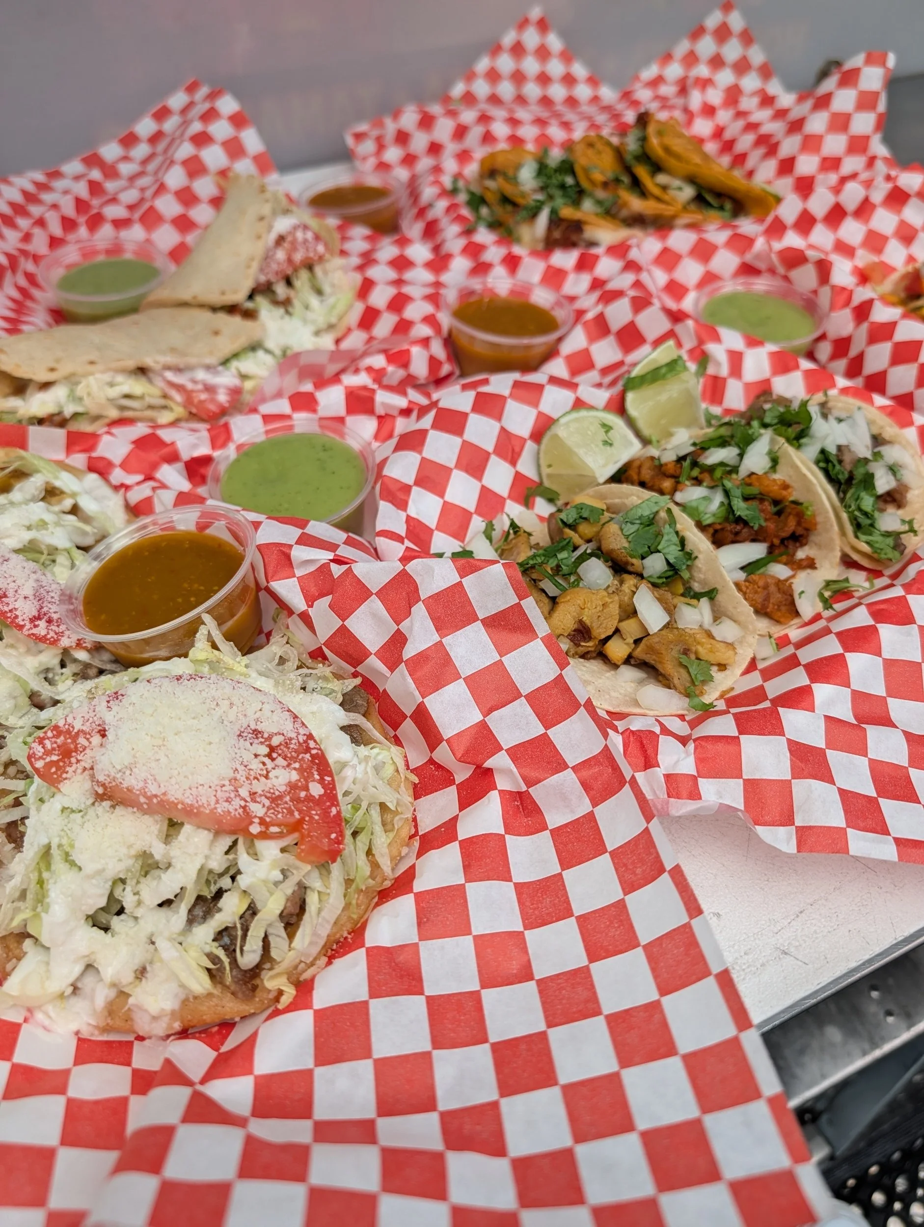 Assorted tacos and a taco salad on red checkered paper with small cups of green and brown sauces.