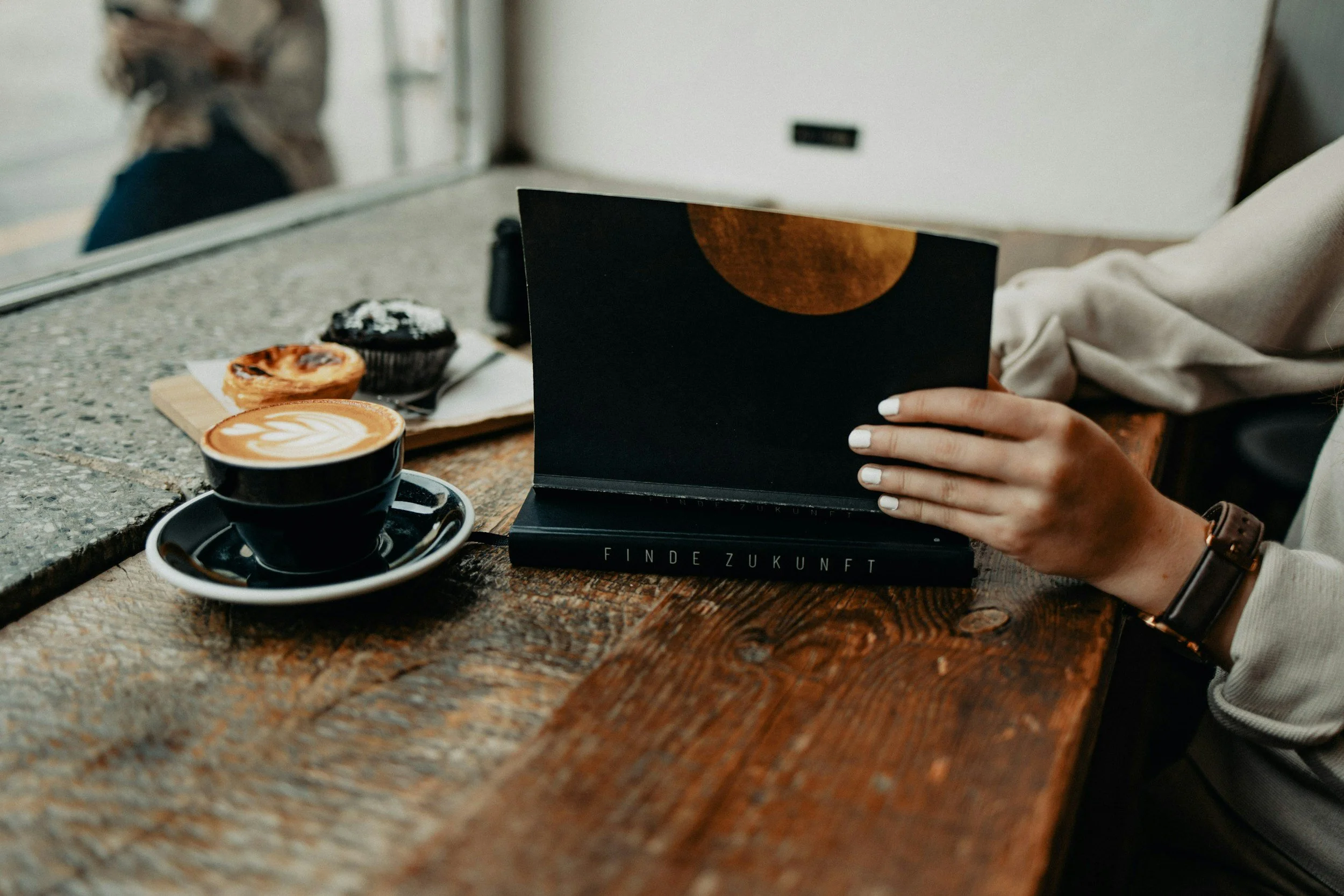 A person sitting at a rustic wooden table in a cafe, holding an open book with a black cover and a moon illustration, with a latte, a muffin, and a pastry on the table.