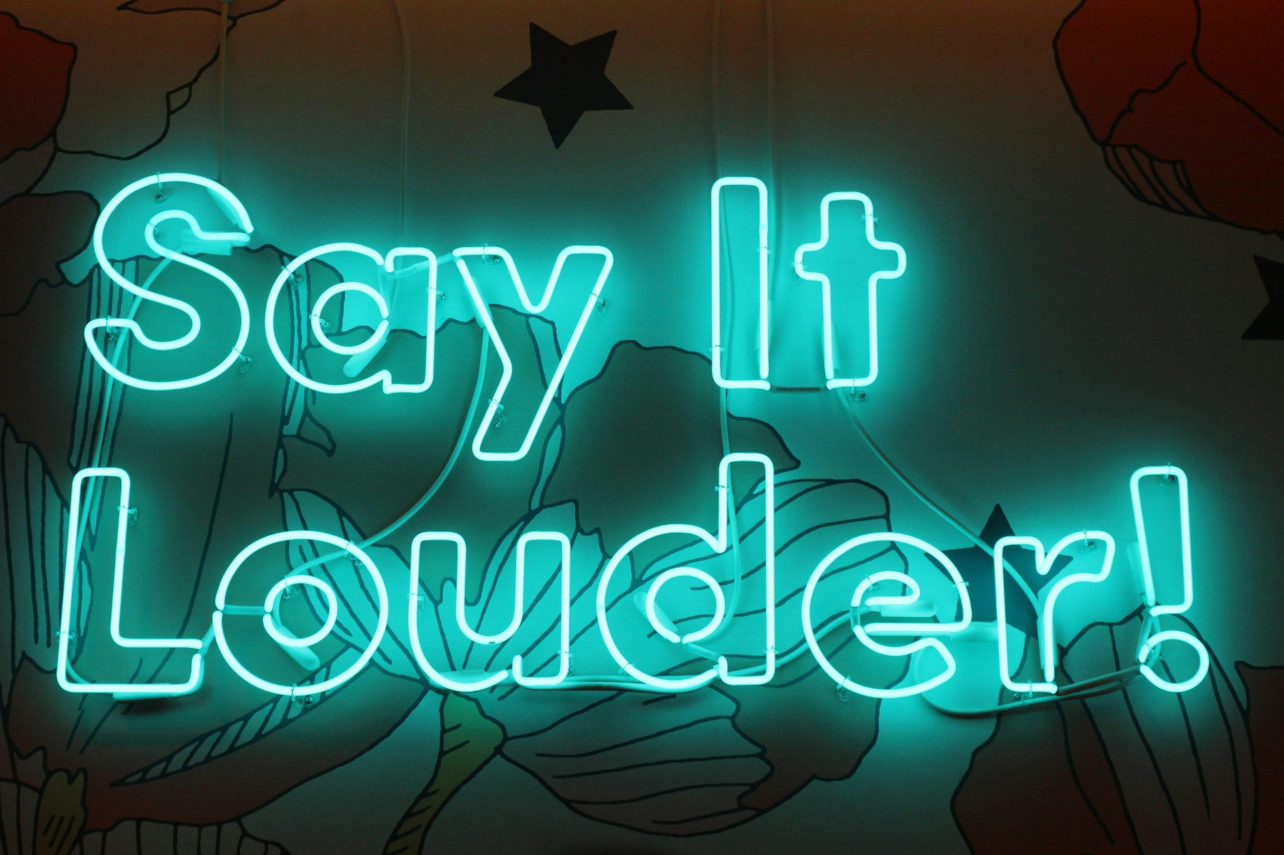 Neon sign that reads 'Say it Louder!' against a colorful wall with floral and star patterns. allow thoughts for mental health
