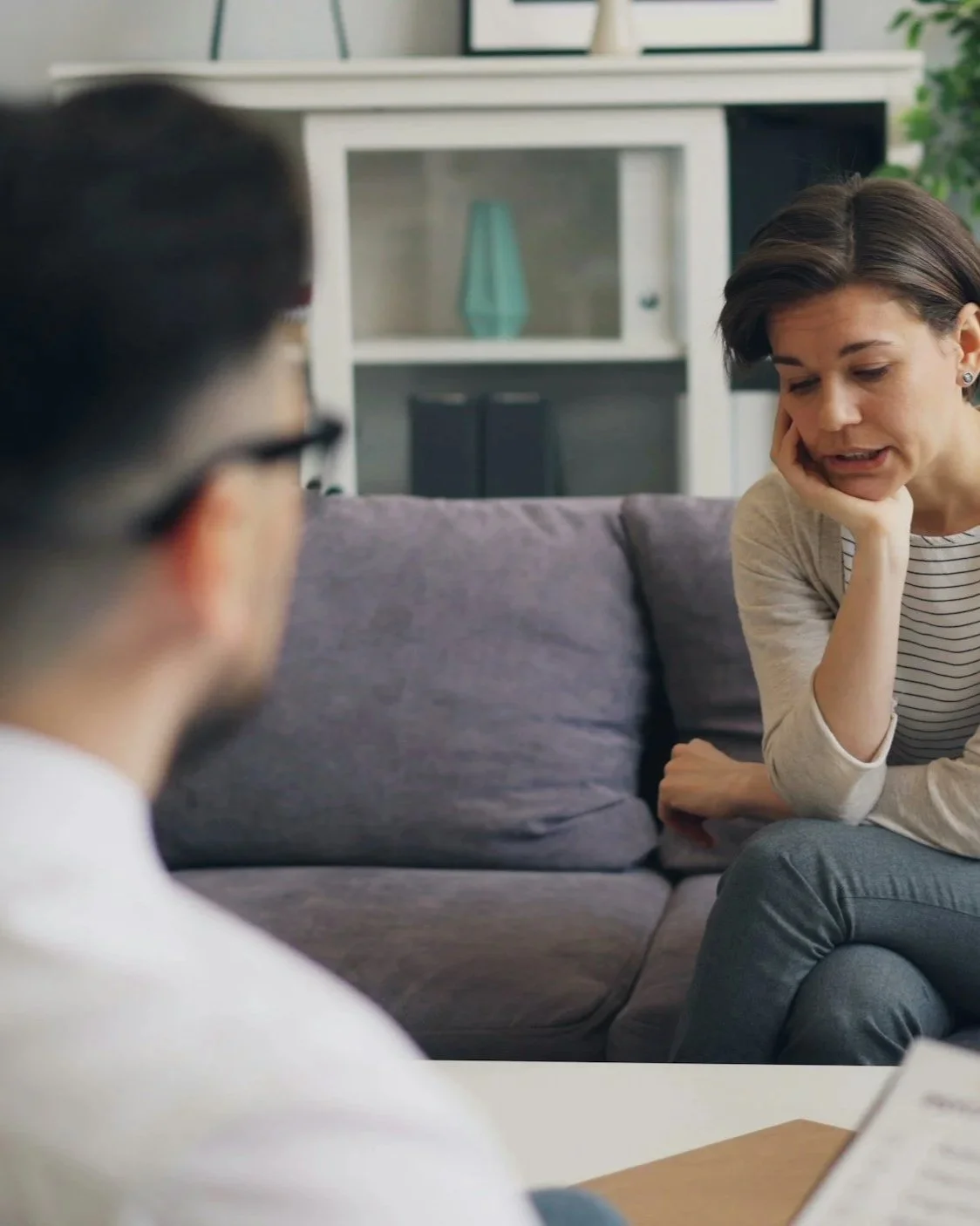 A woman sitting on a couch appears to be upset or distressed, while a person with glasses in the foreground is facing her, possibly in a counseling or therapy session.