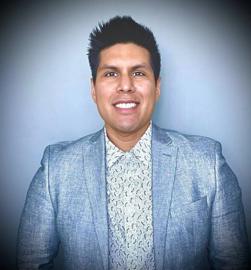 A man in a blue suit and patterned shirt smiling against a plain gray background.