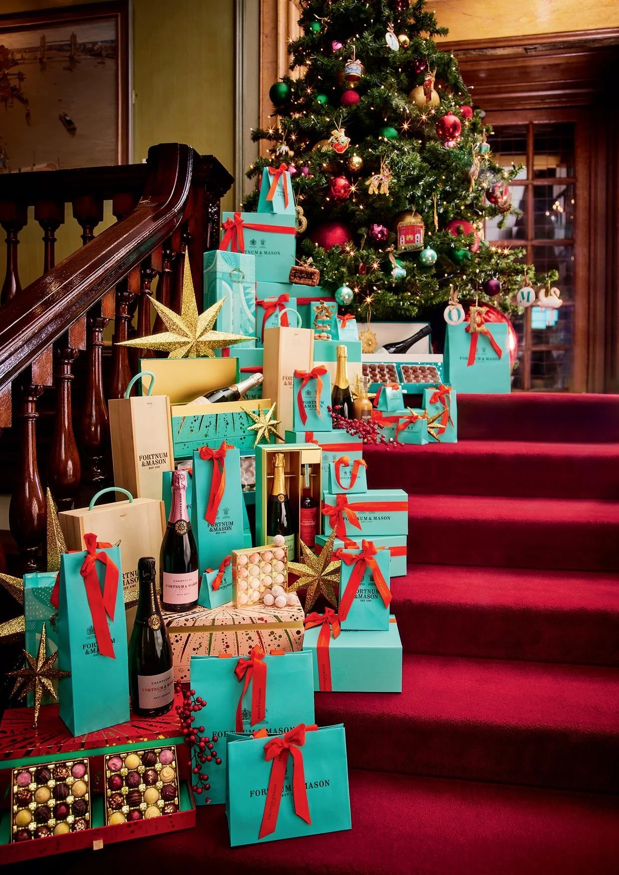 Christmas tree decorated with ornaments and lights, surrounded by blue gift boxes with red ribbons, chocolates, and champagne bottles on a red-carpeted staircase. Fortnum and Mason.