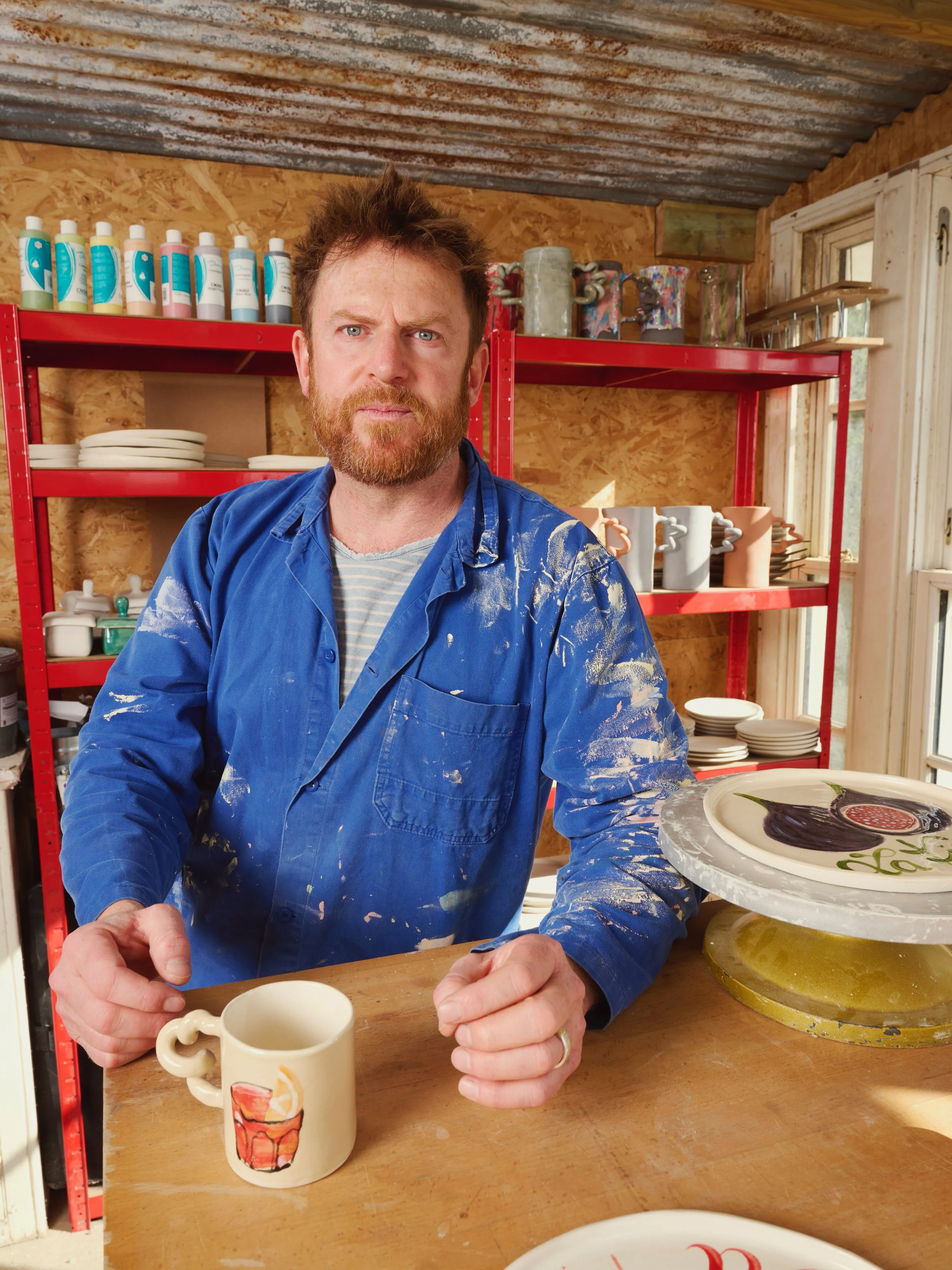 A man with a beard and blue shirt standing in a pottery studio, holding a painted mug, surrounded by pottery and art supplies.