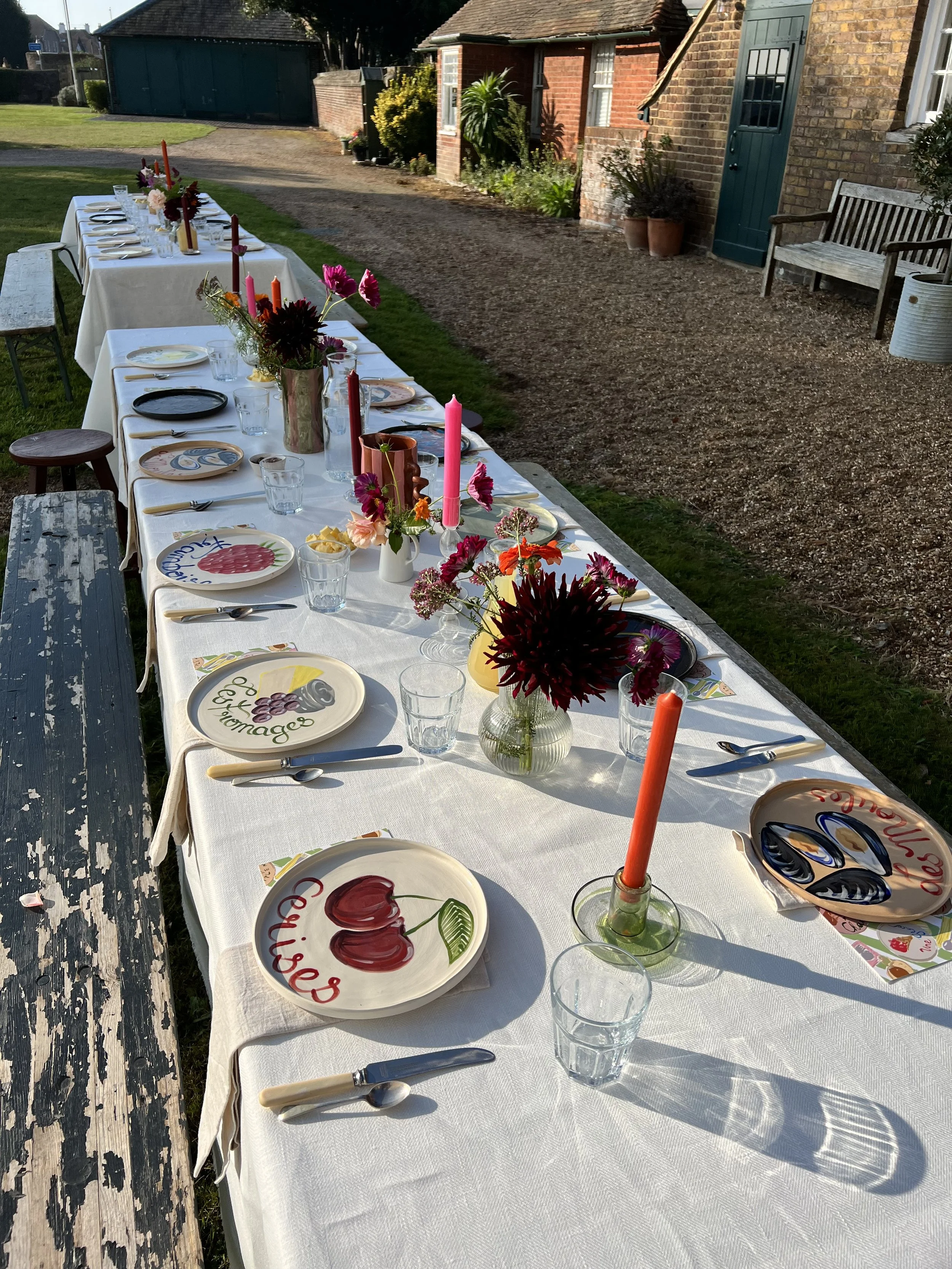 Long outdoor dining table set with white tablecloth, colorful painted plates, clear glasses, silverware, and vibrant floral centerpieces with pink and orange candles. The table is situated in a garden area with a rustic brick building, a bench, and a gravel path nearby.