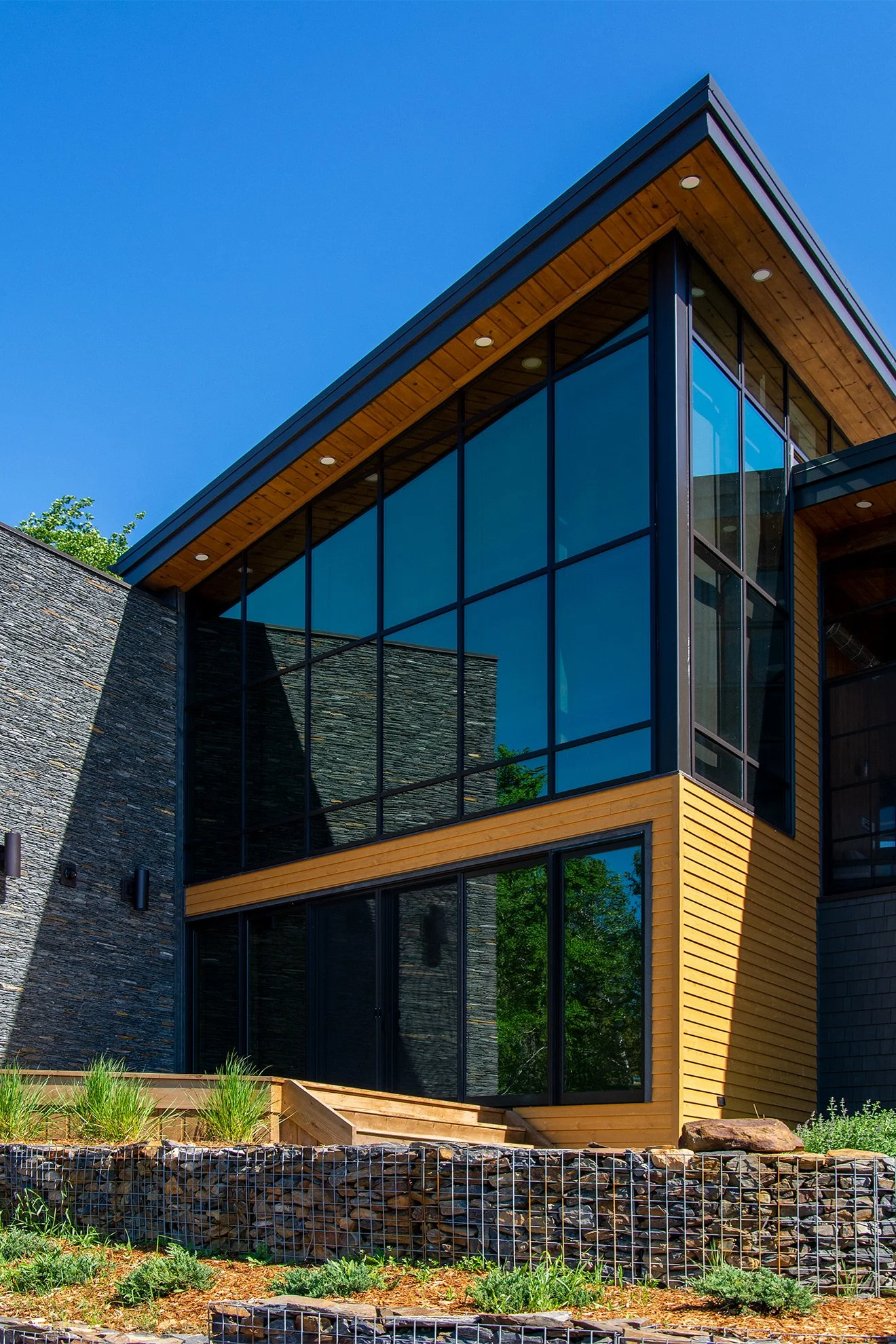 Modern house with large glass windows, wooden siding, and stone accents under a clear blue sky.