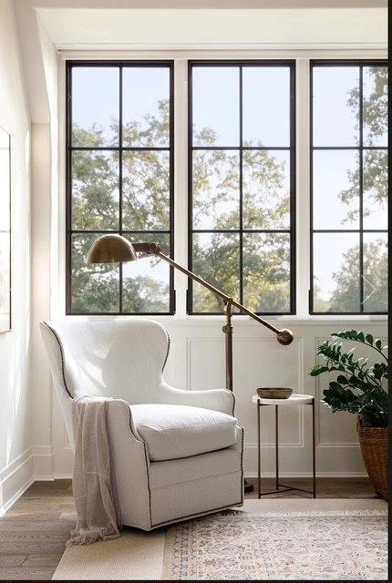 A white armchair with a blanket draped over its side, next to a small table with a plant, near a large window with multiple panes and a view of trees outside.