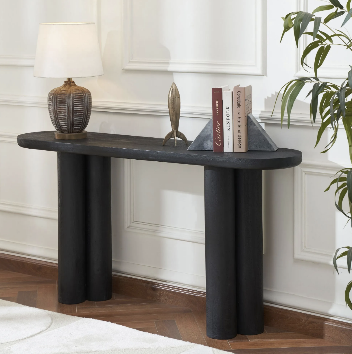Black console table with decorative lamp, metallic rocket, and stack of books with geometric bookends, against a white paneled wall.