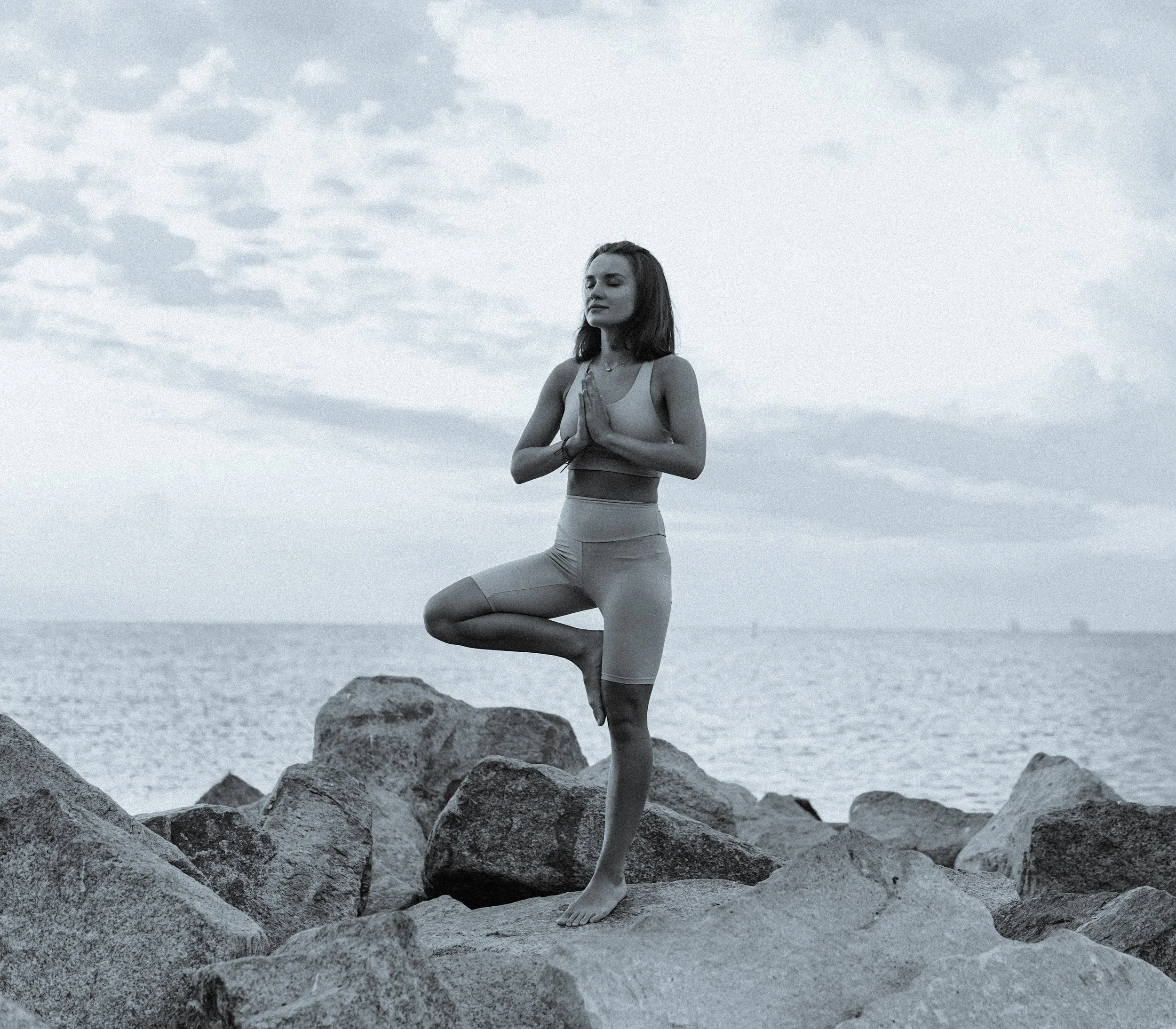 Woman practicing yoga tree pose on coastal rocks by the ocean at sunrise – Eternal Now Yoga, Estero Florida.