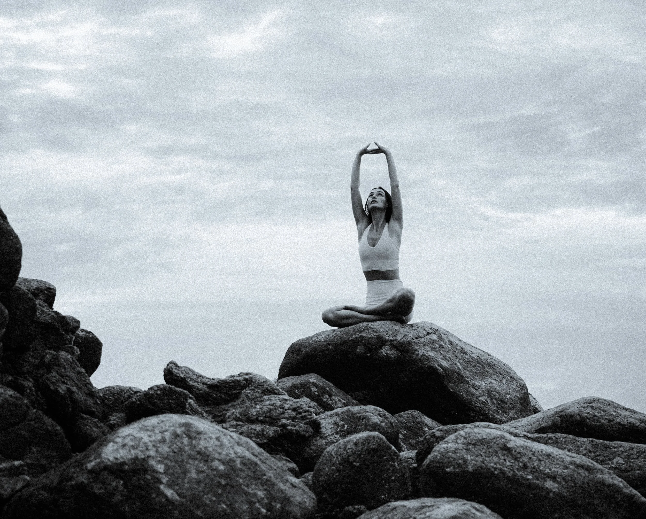 Woman practicing yoga on seaside rocks at sunrise, seated in lotus pose with arms raised – Eternal Now Yoga, Estero Florida.