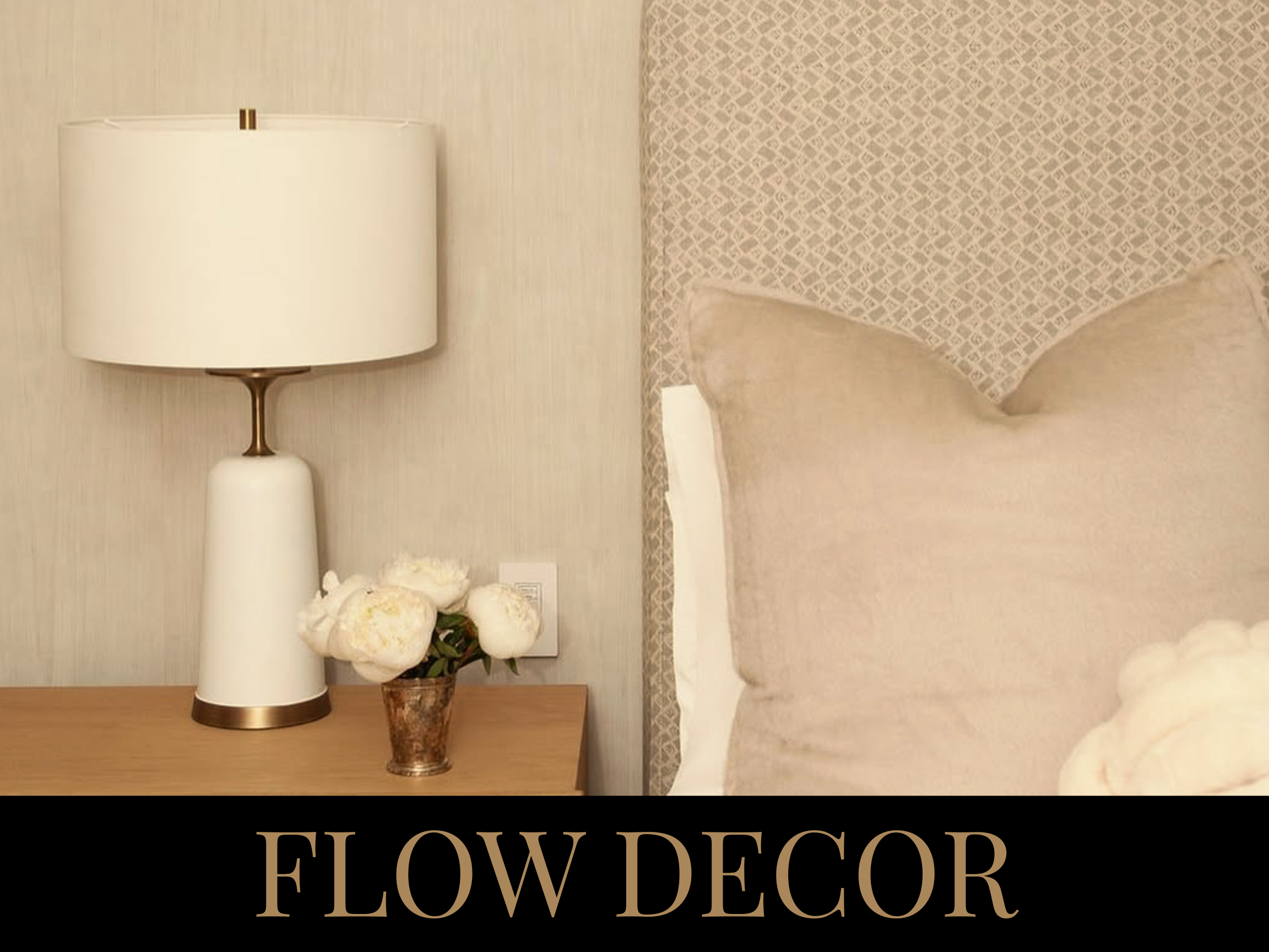 A bedside table with a white lamp and a vase of white flowers, next to a bed with beige pillows and a patterned headboard.