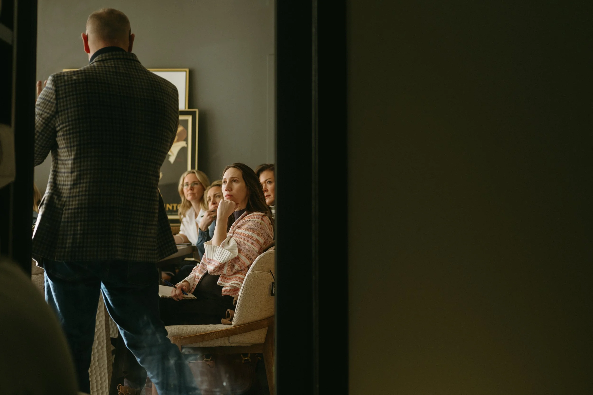 A group of women sitting at a conference or meeting, listening to a speaker standing in front of them, with the speaker facing away from the camera.