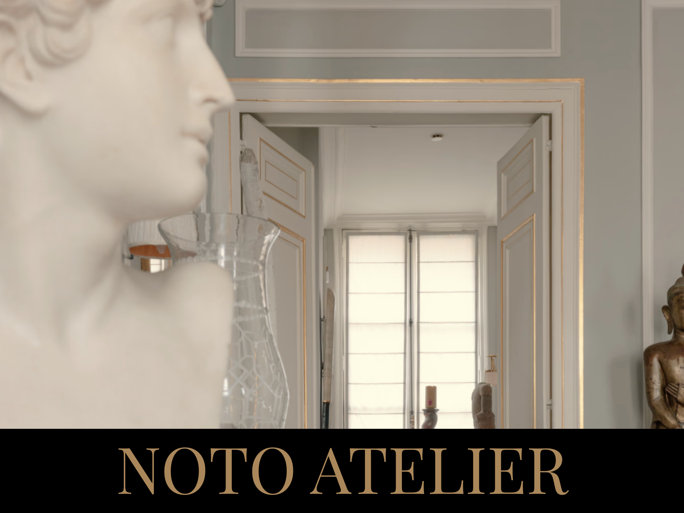 Interior view of a room with classical decor, featuring a white bust sculpture, a glass vase, and a doorway with double doors leading to a brightly lit space with large windows.