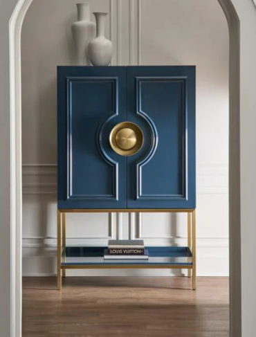 A blue cabinet with gold accents and two doors, placed on a gold stand with a lower shelf holding books, in a room with beige walls, white decorative vases on top, and hardwood flooring.