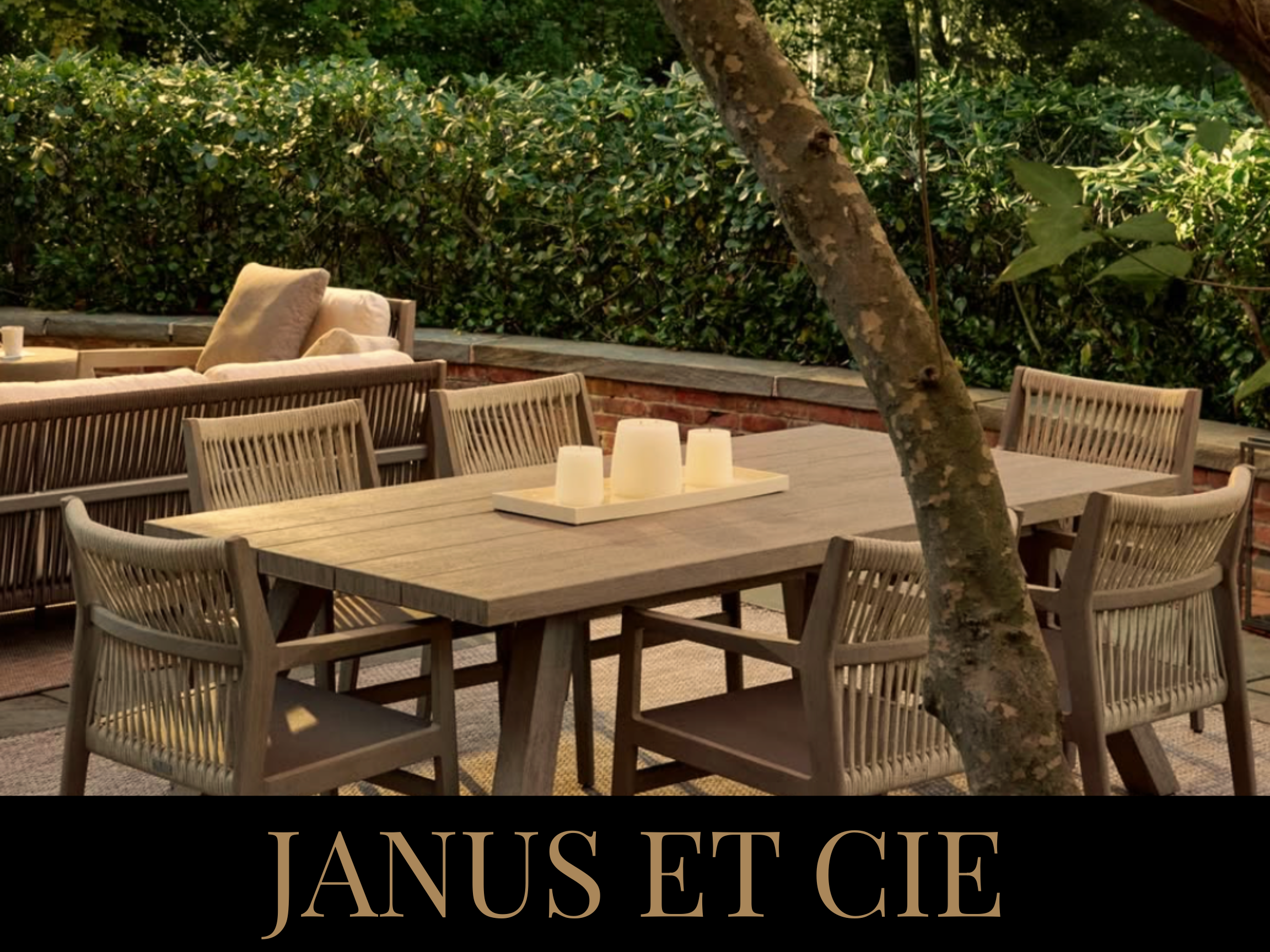 An outdoor patio with a wooden dining table, six matching chairs, a tray with white candles, a cushioned sofa with pillows, a long wooden railing, dense green foliage, and a tree trunk in the foreground. Text overlay reads "JANUS ET CIE".