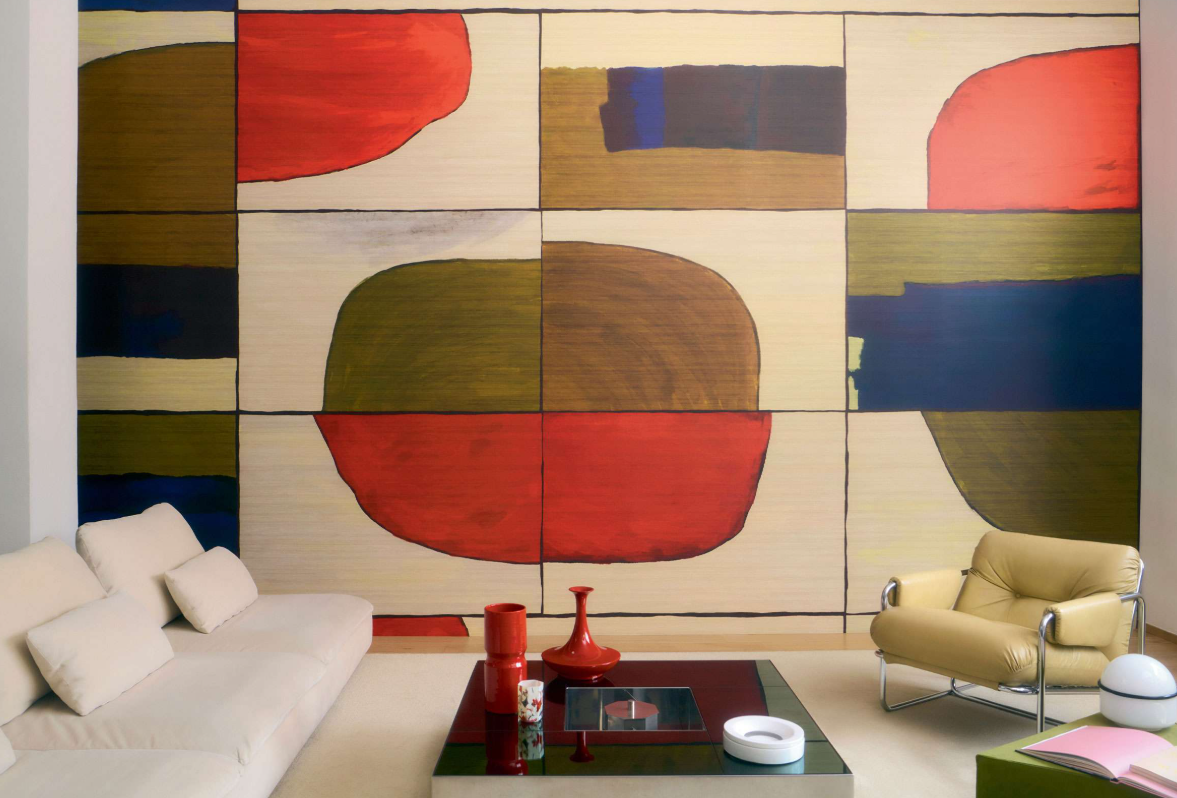 Modern living room with a large abstract art wall mural featuring geometric shapes in red, brown, black, blue, and cream. The room includes a beige sofa, a cream armchair, a black glass-top table with red and white vases, and a green ottoman with a pink book on top.
