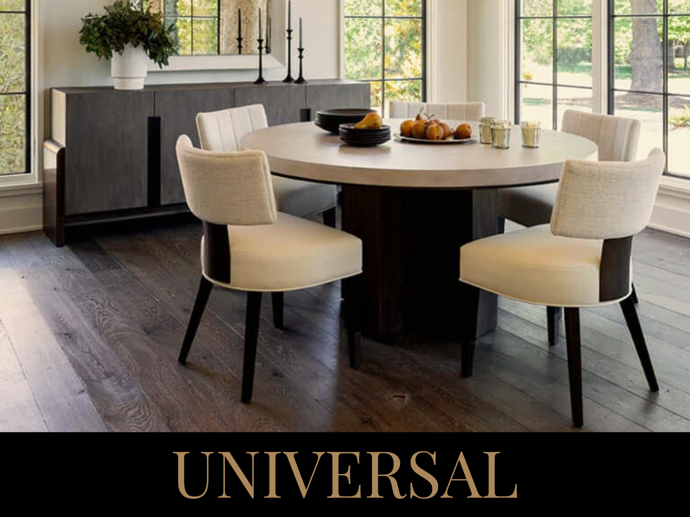 A round dining table with six beige upholstered chairs, set with some fruit and candles in a bright room with large windows and wooden floors.