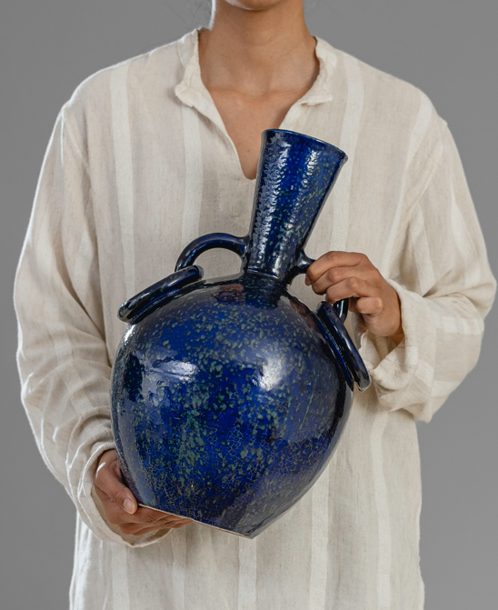 Person holding a large blue ceramic vase with a narrow neck against a gray background.